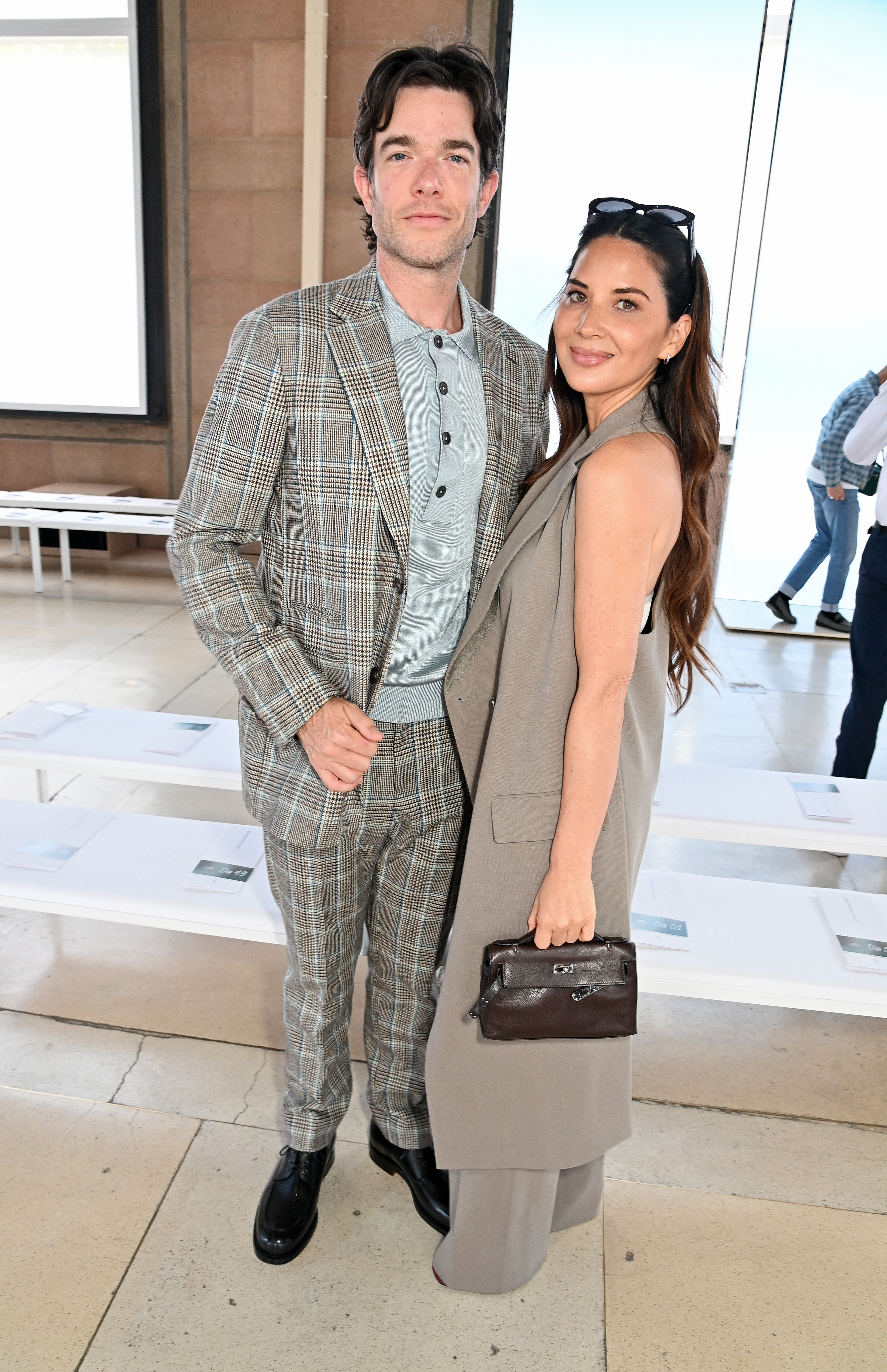 John Mulaney in a plaid suit and Olivia Munn in a long vest pose together at an event