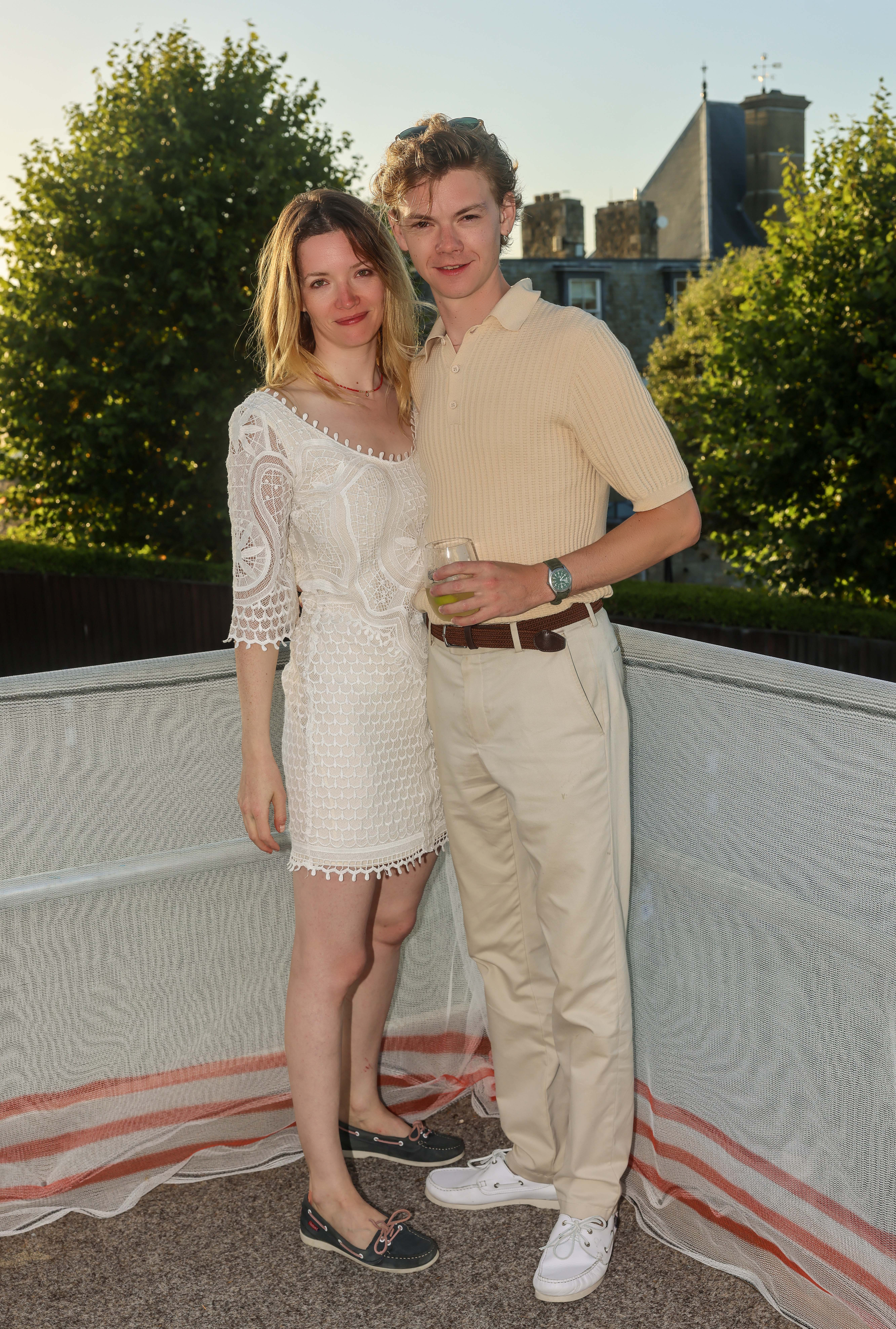 Talulah Riley and Thomas Brodie-Sangster stand together outdoors. Riley wears a lace dress and flats, Brodie-Sangster in a polo shirt and slacks holding a drink