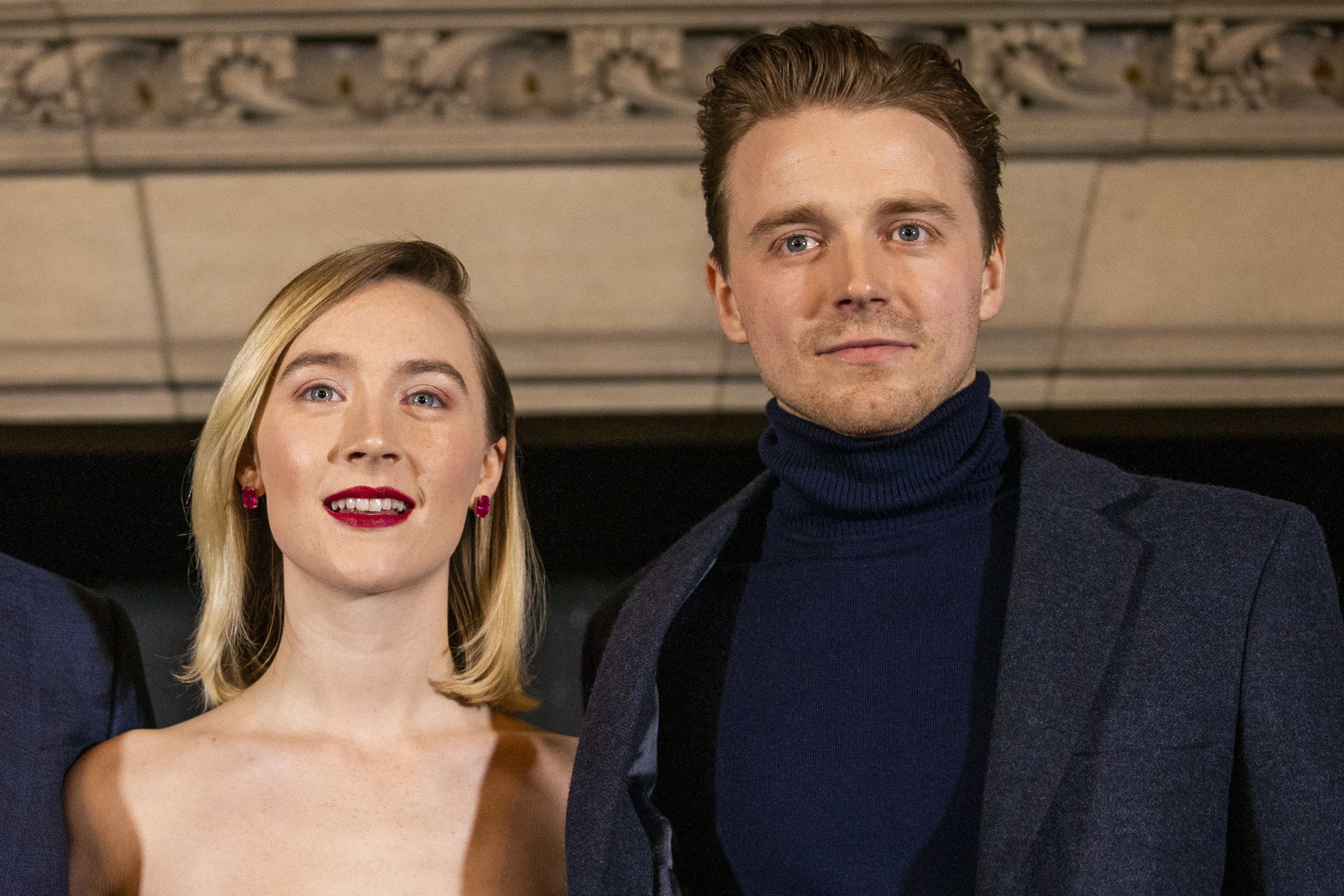 Saoirse Ronan and Jack Lowden pose together, she is in a strapless dress with red earrings, and he wears a dark suit with a turtleneck underneath