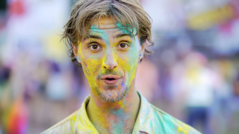 Person with colorful paint on face and hair, looking surprised, likely participating in a Holi festival or similar event