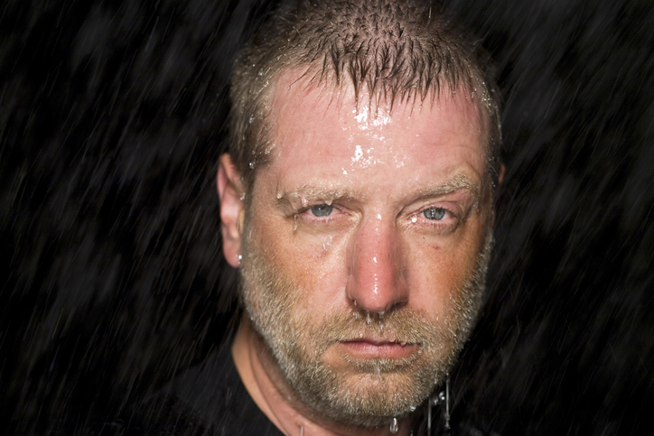 A close-up of a solemn-looking man with short, wet hair and a beard, likely standing in rain or water, with water droplets on his face