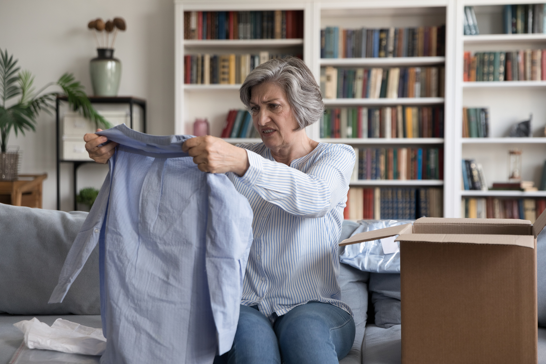 An older woman with short gray hair, wearing a casual blouse, looks dissatisfied while holding up a shirt from an open shipping box in her living room