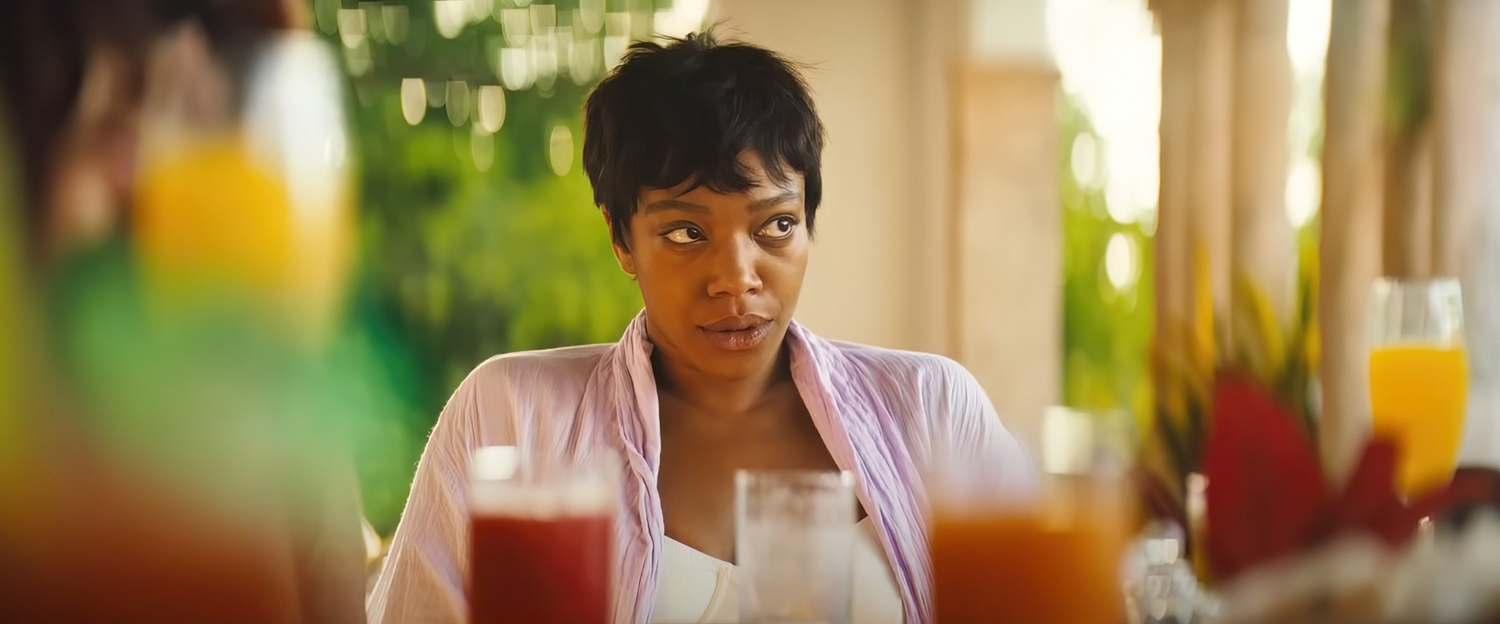 Naomi Ackie is seated at an outdoor event, looking off to the side with a slight smile. She is wearing a light-colored outfit