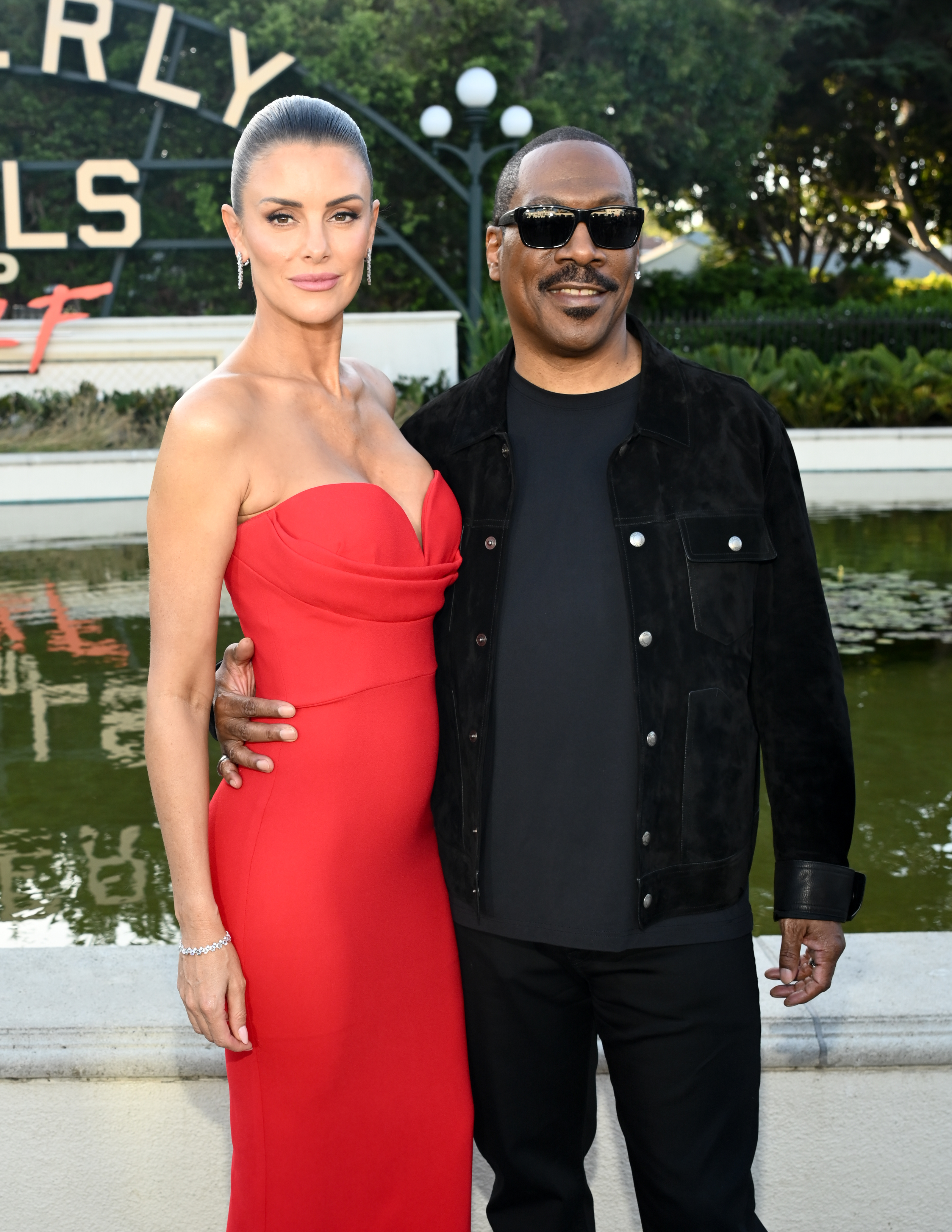 Eddie Murphy in a jacket and sunglasses stands with Paige Butcher in a strapless gown, posing outdoors in front of a sign and garden