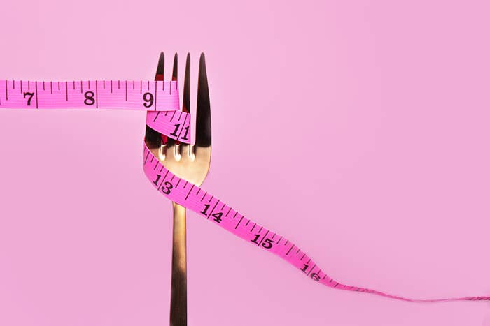 A silver fork with a pink measuring tape wrapped around its prongs against a pink background, symbolizing dieting or portion control