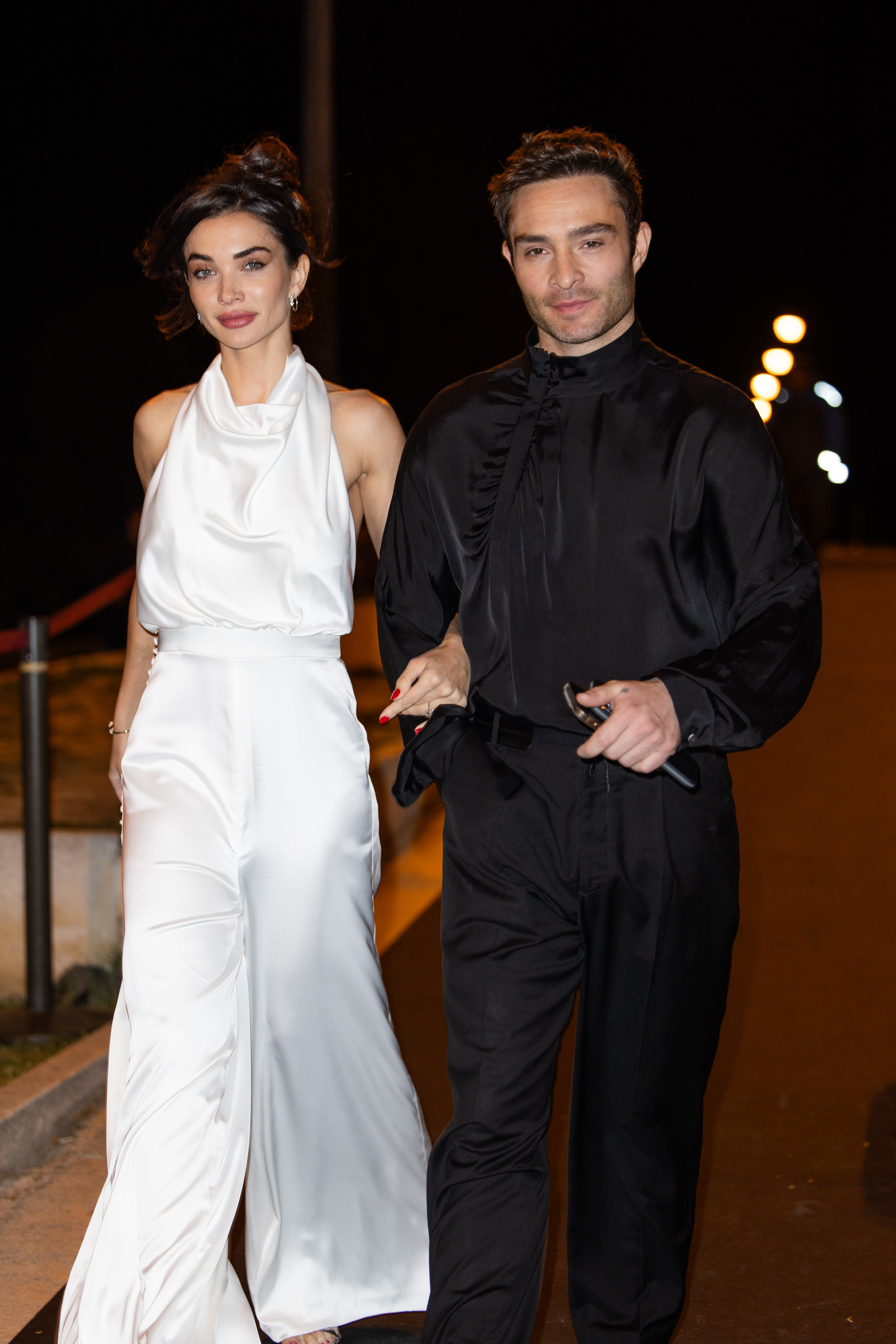 Amy in a sleeveless jumpsuit and Ed in a high-collared shirt and pants, walking at night