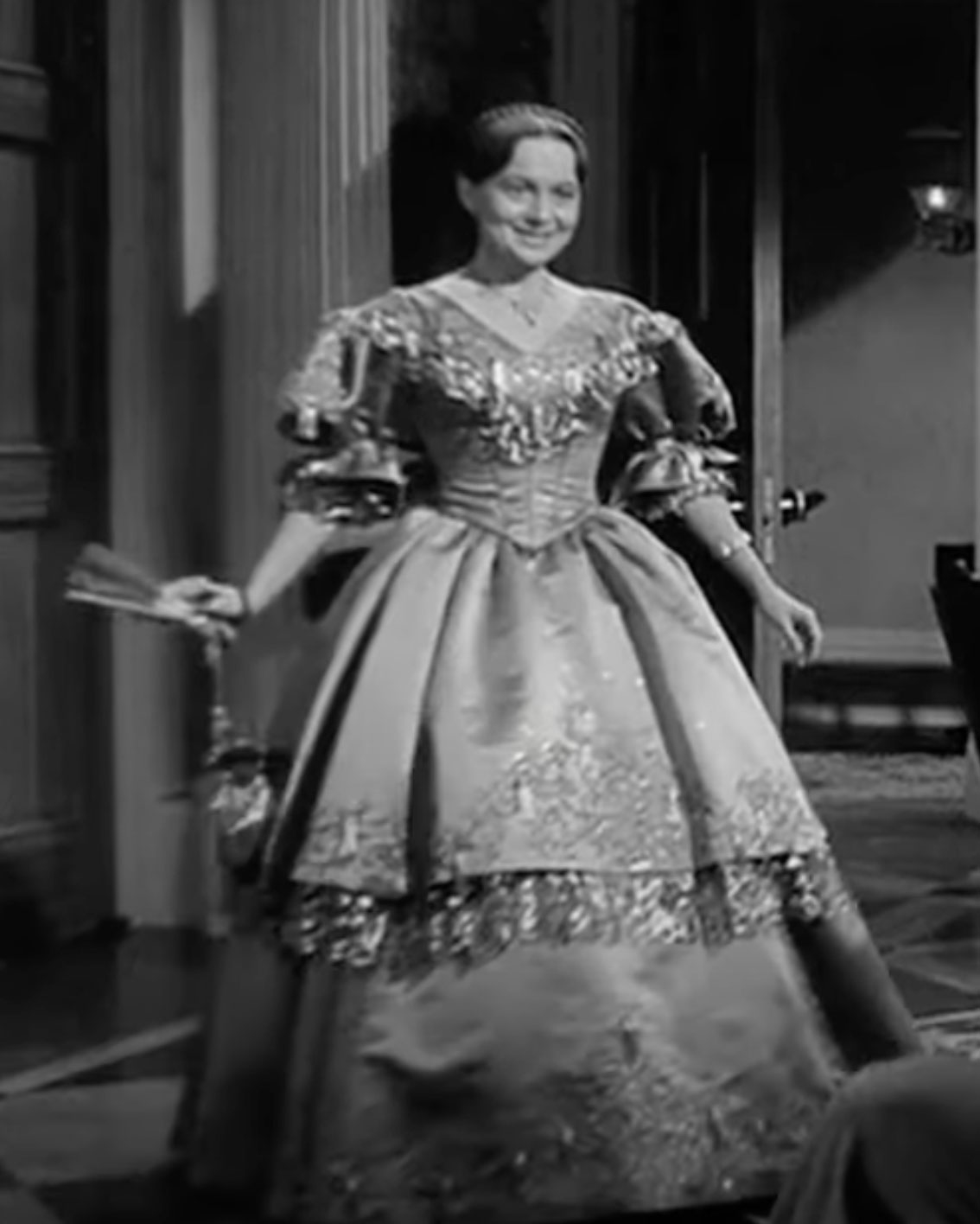 Unknown actress in an elegant, voluminous 19th-century style gown featuring ornate detailing, holding a fan, and standing in a lavishly decorated room