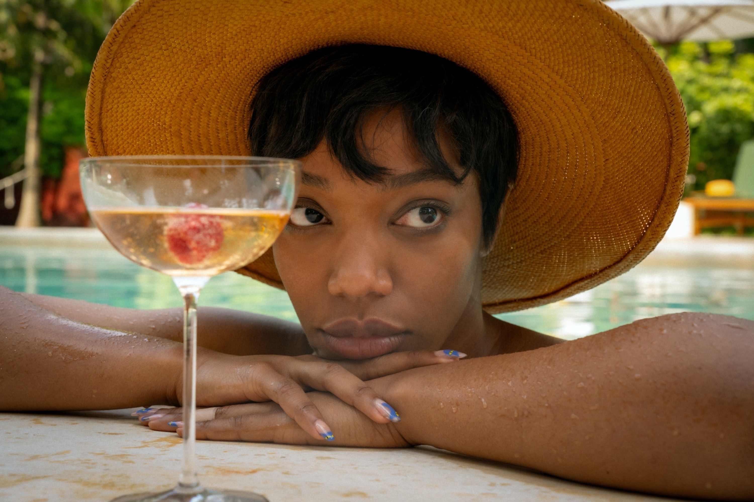 Naomi Ackie wearing a wide-brimmed hat, resting her arms on the edge of a pool, a glass with a drink and a cherry inside next to her
