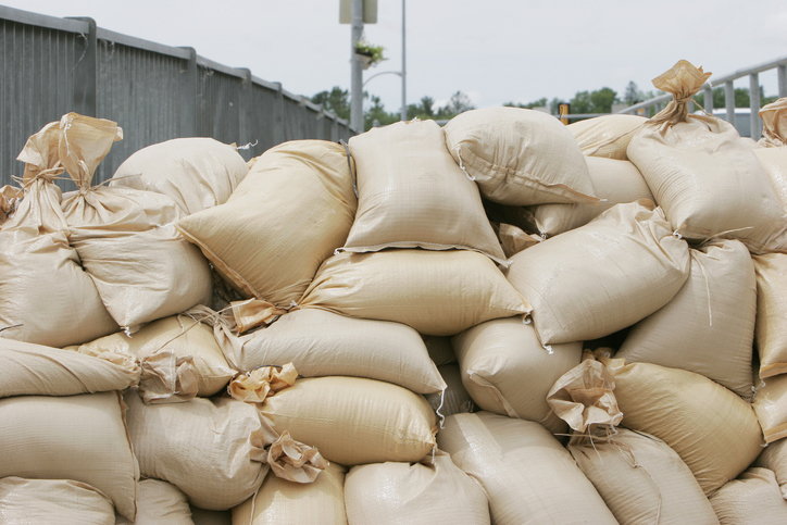 A stack of beige sandbags are piled against a concrete barrier, likely for flood prevention. There is no discernible text or people in the image