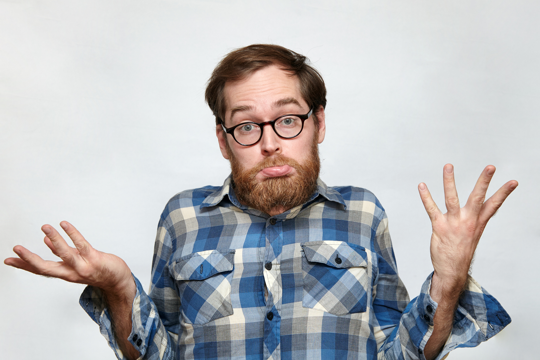 A man with glasses and a beard in a plaid shirt shrugs with a confused expression, raising both hands