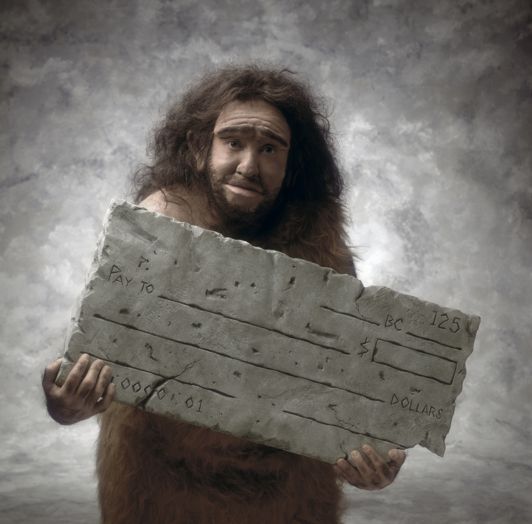 A caveman stands holding a large stone carved to look like a cheque. The cheque is made out for 125 BC dollars, highlighting the contrast between ancient times and modern financial instruments