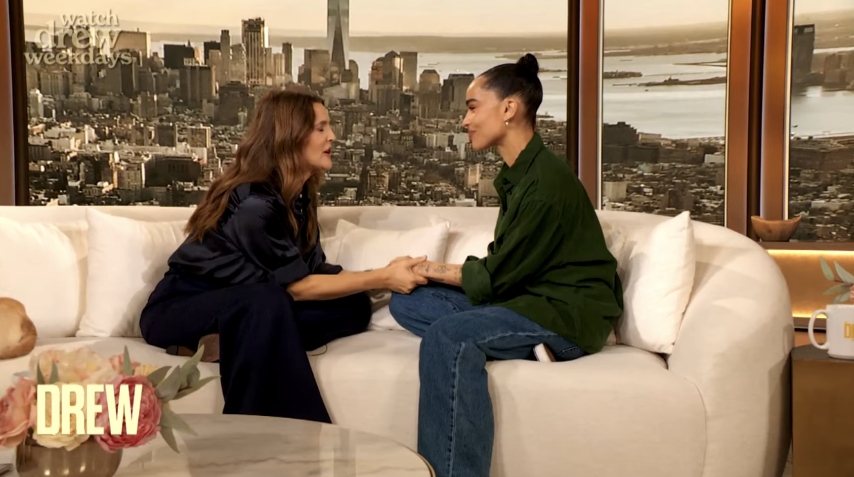 Drew Barrymore and guest sit on a couch holding hands, engaged in conversation on "The Drew Barrymore Show" set with a cityscape backdrop