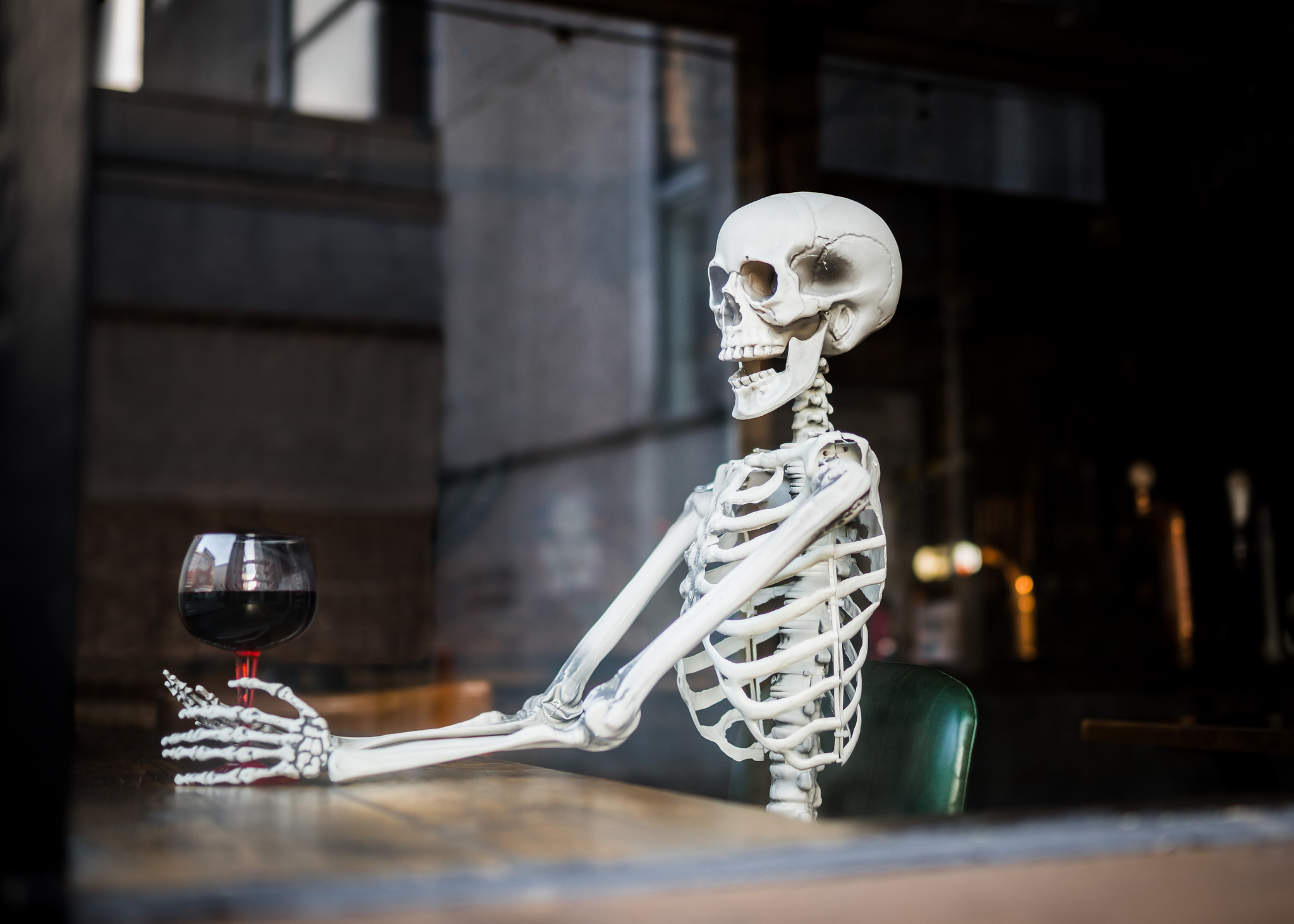 A human skeleton is sitting at a table holding a glass of wine in a humorous, staged setting