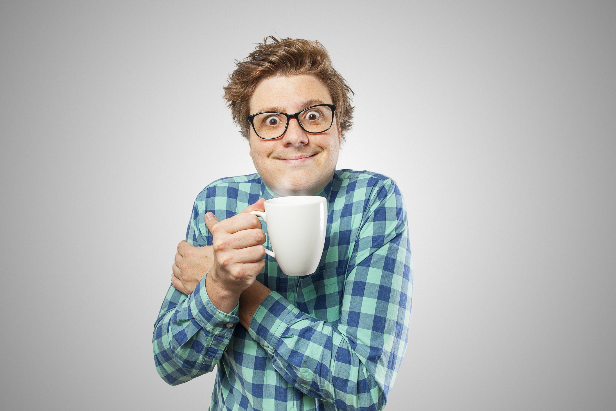 A person with glasses and tousled hair wearing a checkered shirt holds a mug close to their chest with a wide-eyed, excited expression