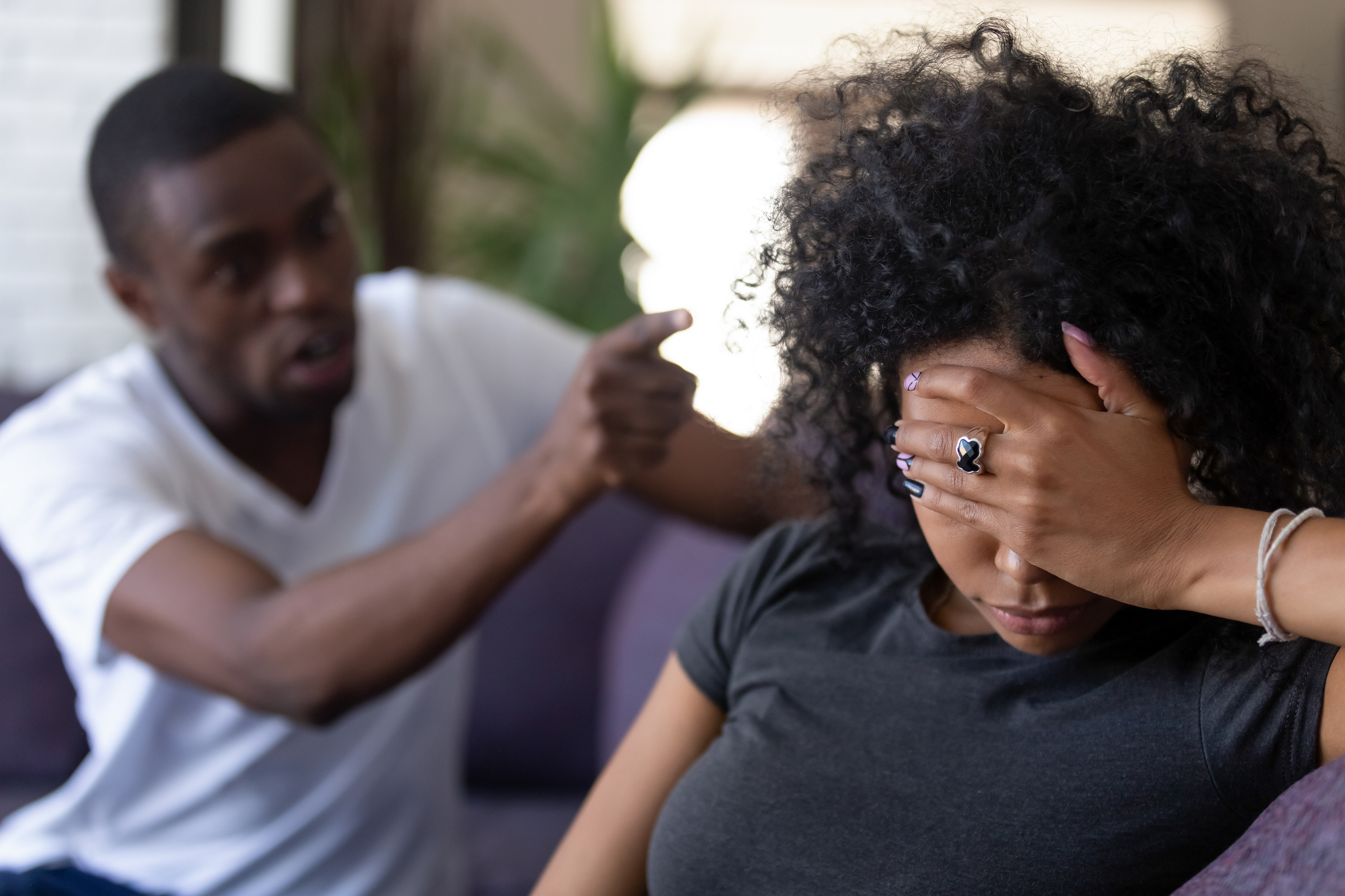 A man, visibly upset, points his finger while speaking to a woman sitting on a couch with her head in her hand, appearing distressed