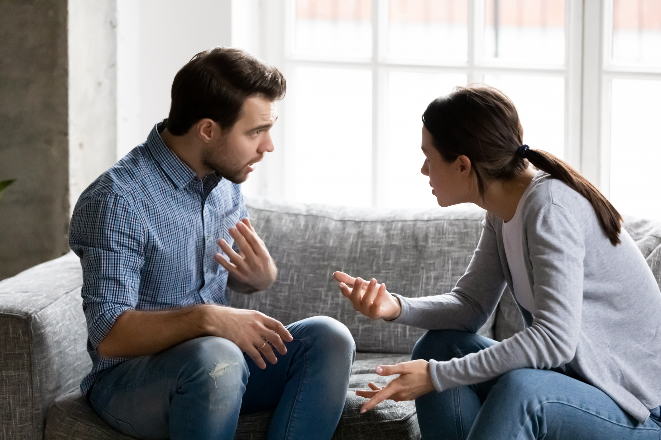 A man and woman sit on a couch having an intense conversation, gesturing with their hands, appearing to be in a disagreement