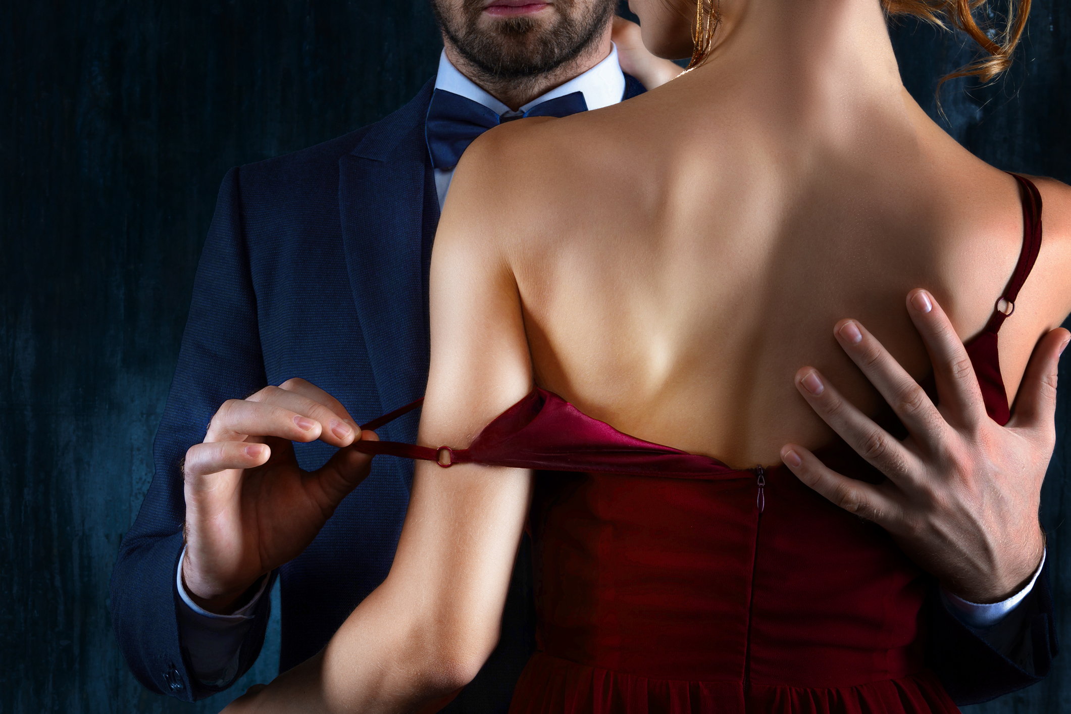 A person in a suit gently pulls down the strap of another person's elegant dress, creating an intimate moment