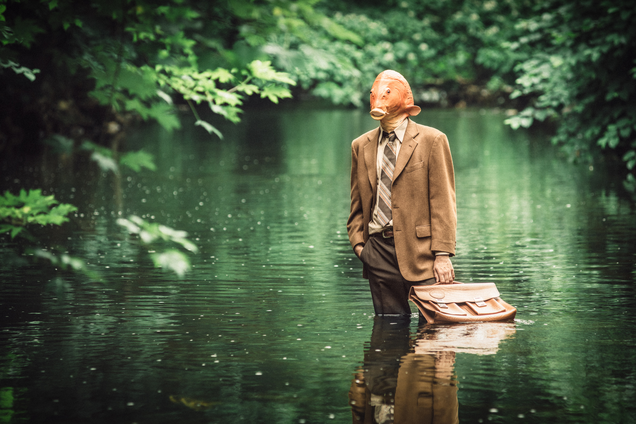 A person in a gas mask and a formal suit stands in a forest pond holding a briefcase