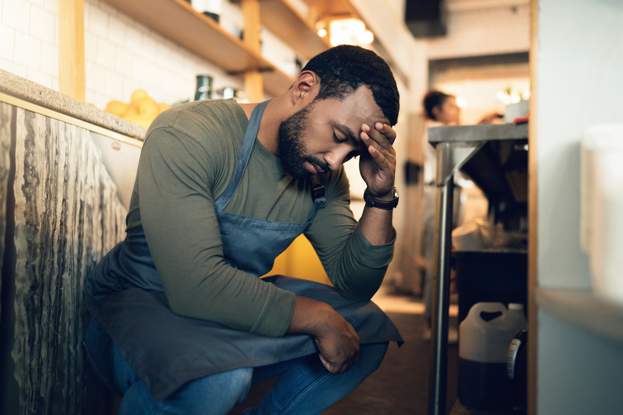 A person in a kitchen, wearing an apron, kneels down and holds their head with a troubled expression. Kitchen equipment and shelves are visible in the background