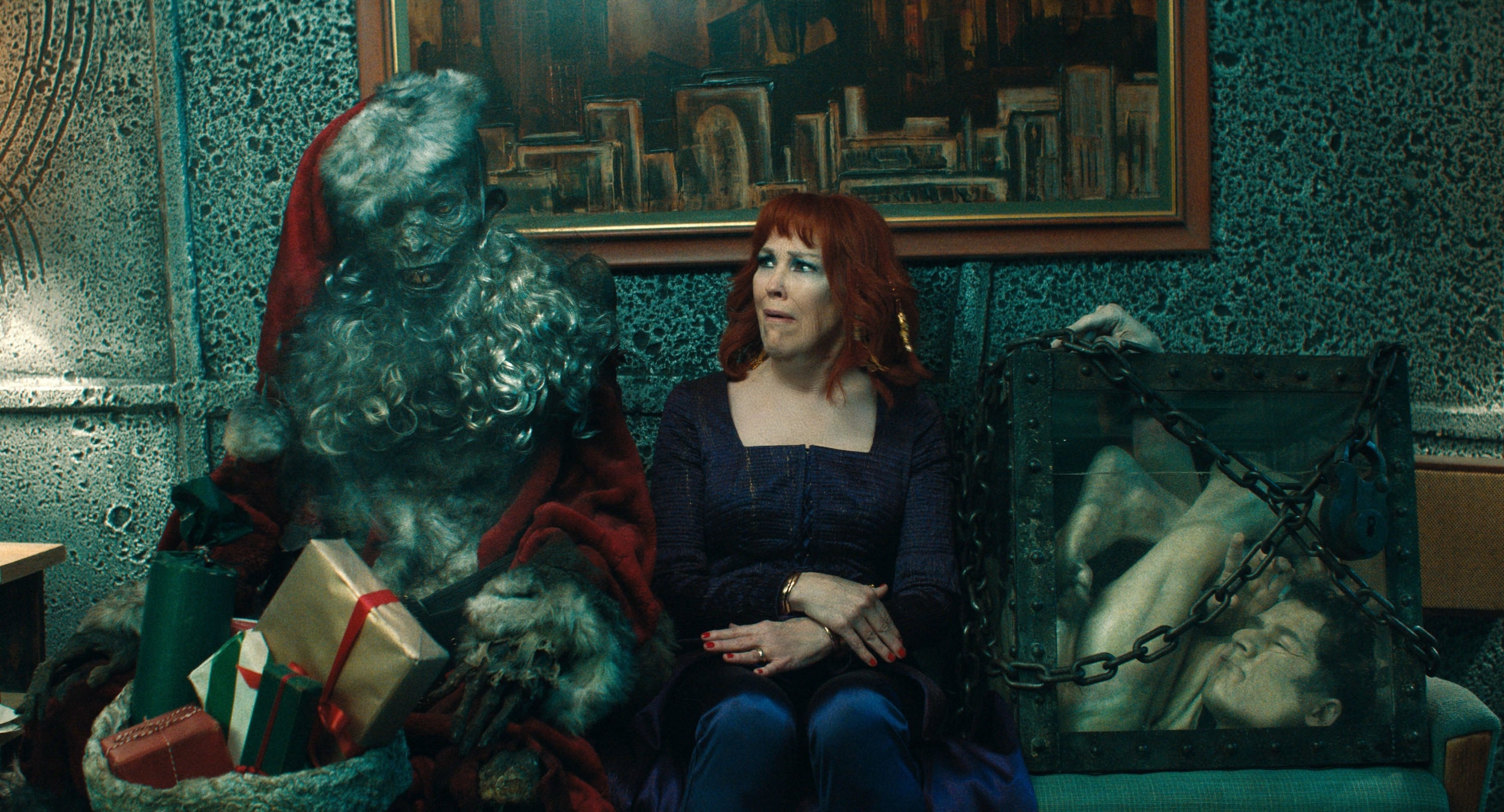 Image of a scene from the TV show "What We Do in the Shadows." Natasia Demetriou sits on a couch next to a ghastly Santa Claus-like figure holding gifts
