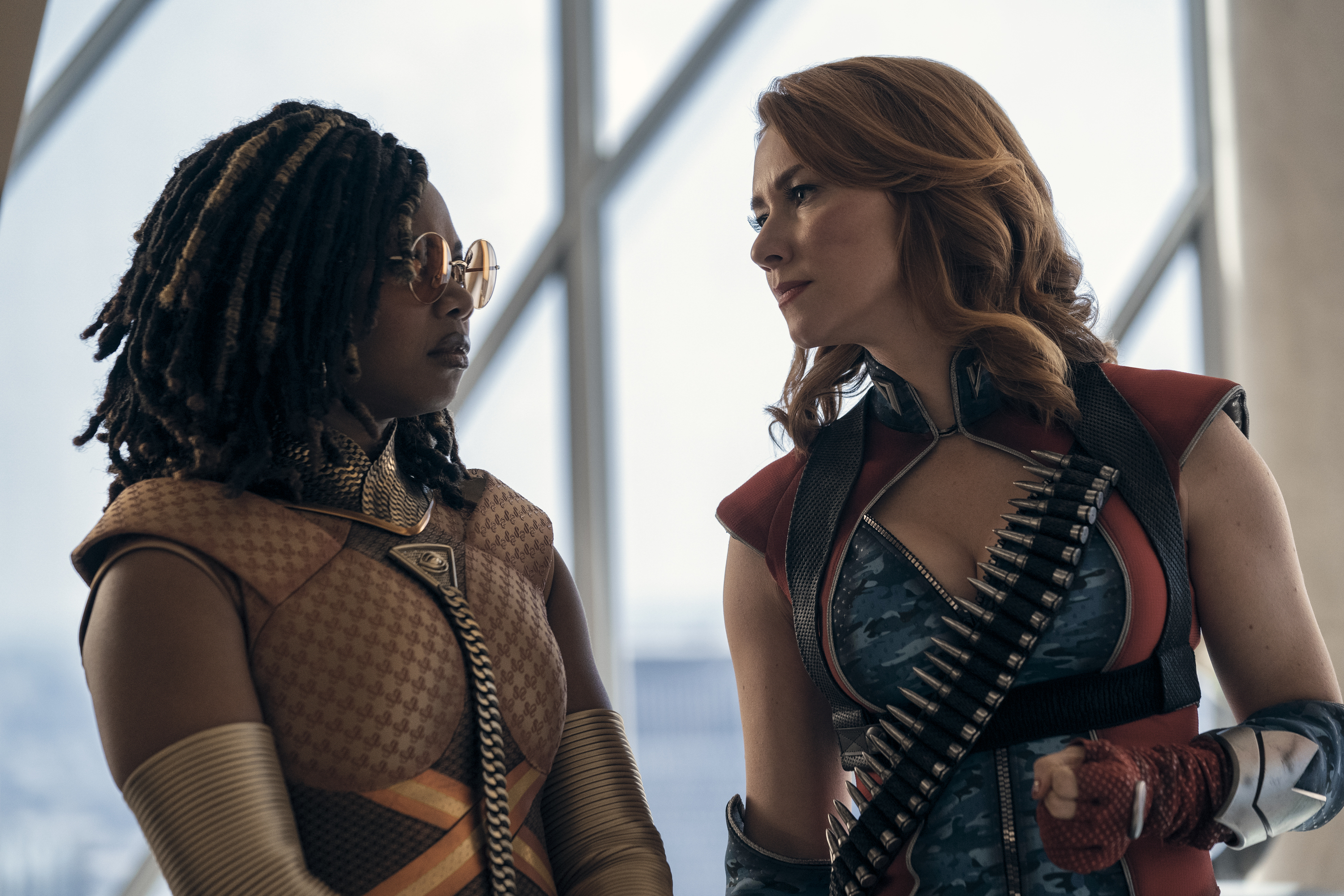 Sister Sage and Firecracker in superhero costumes, engaged in conversation indoors