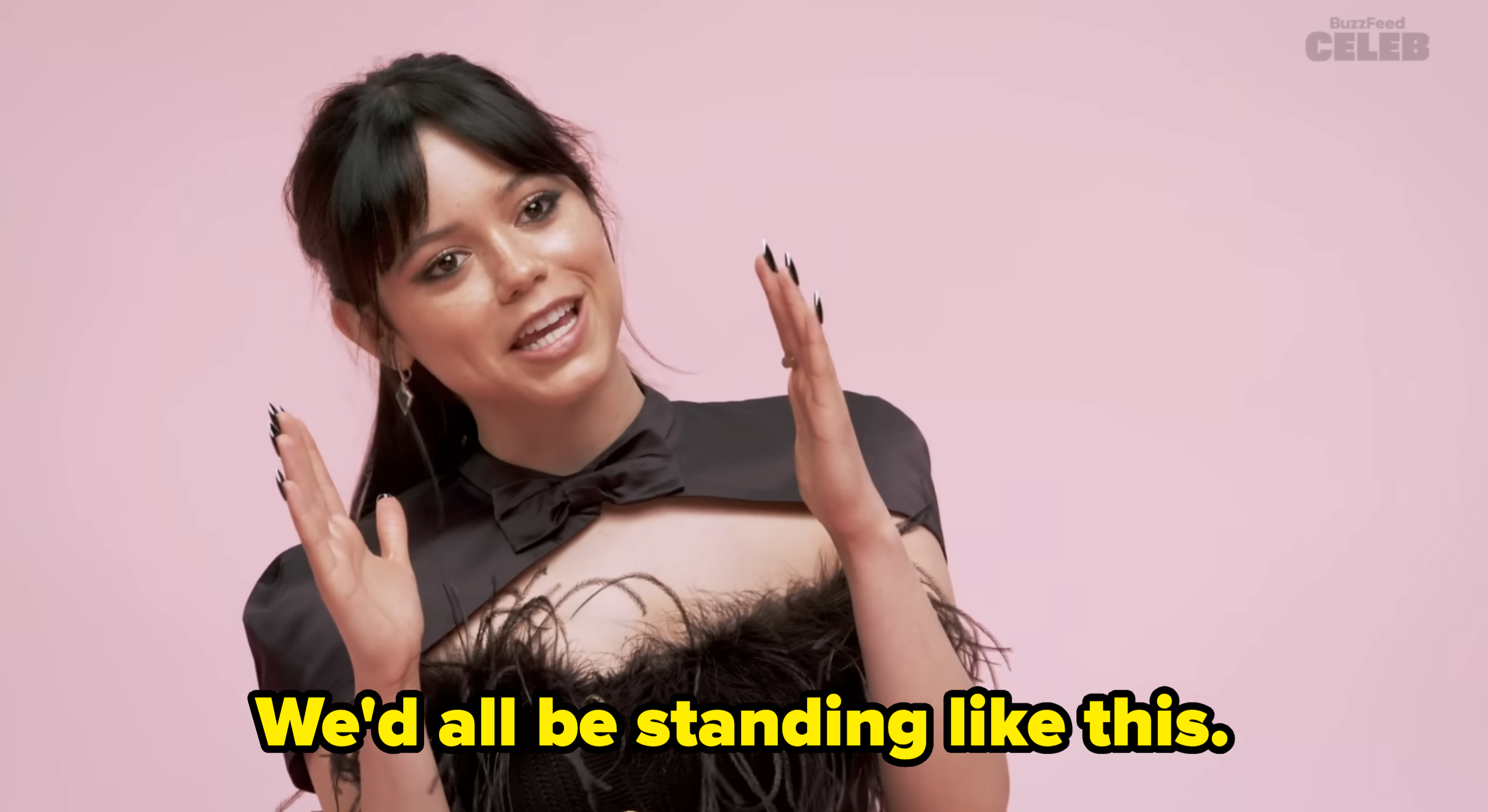 Jenna Ortega wearing a black frilled outfit and cape, gesturing animatedly, in a pastel background for a BuzzFeed Celeb segment