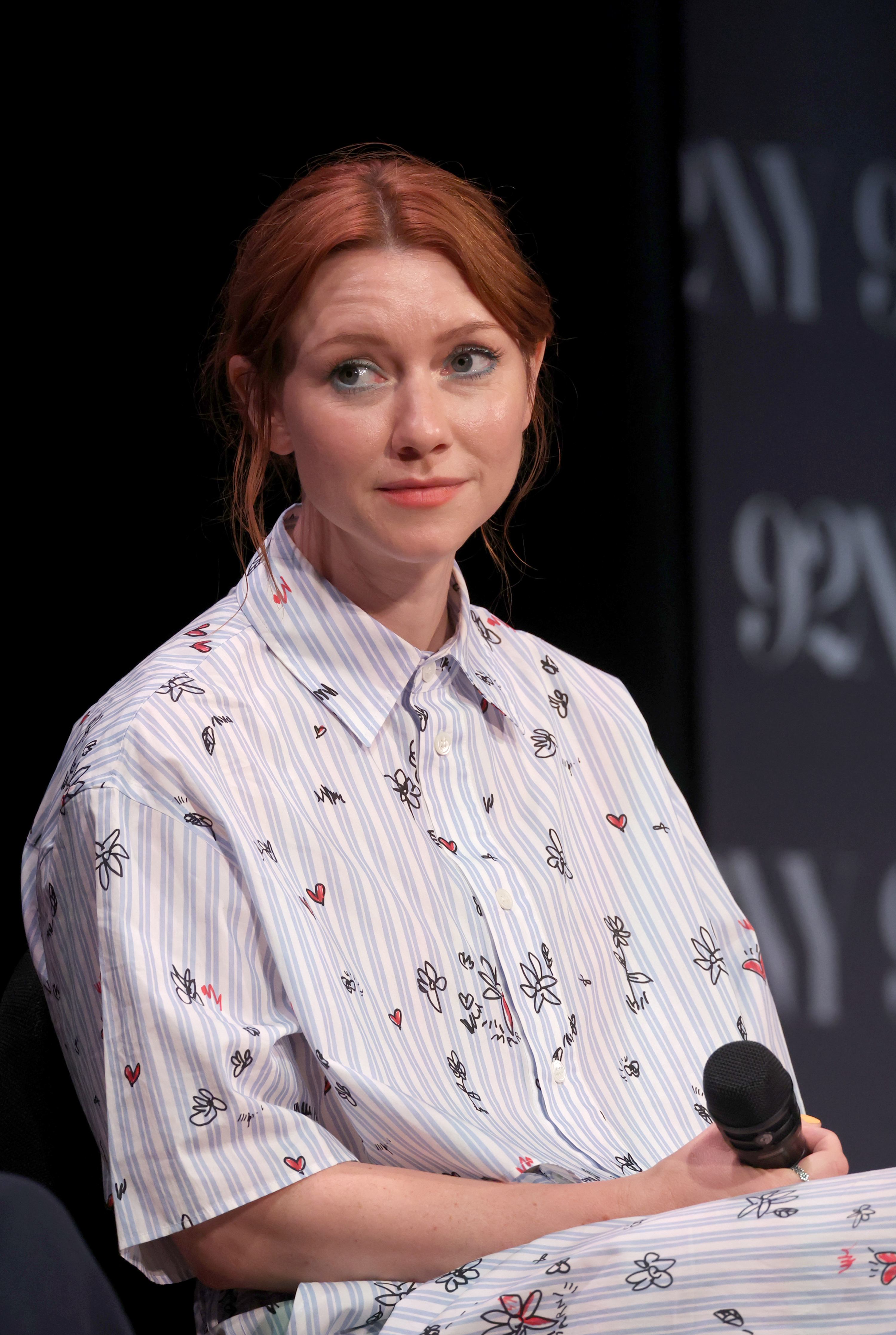 Valorie Curry at an event, wearing a patterned button-up shirt and holding a microphone