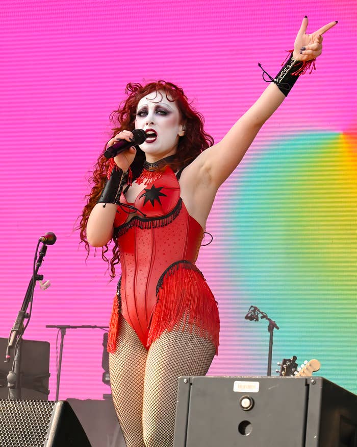 Performer in elaborate red stage outfit gestures energetically while holding a microphone during a concert