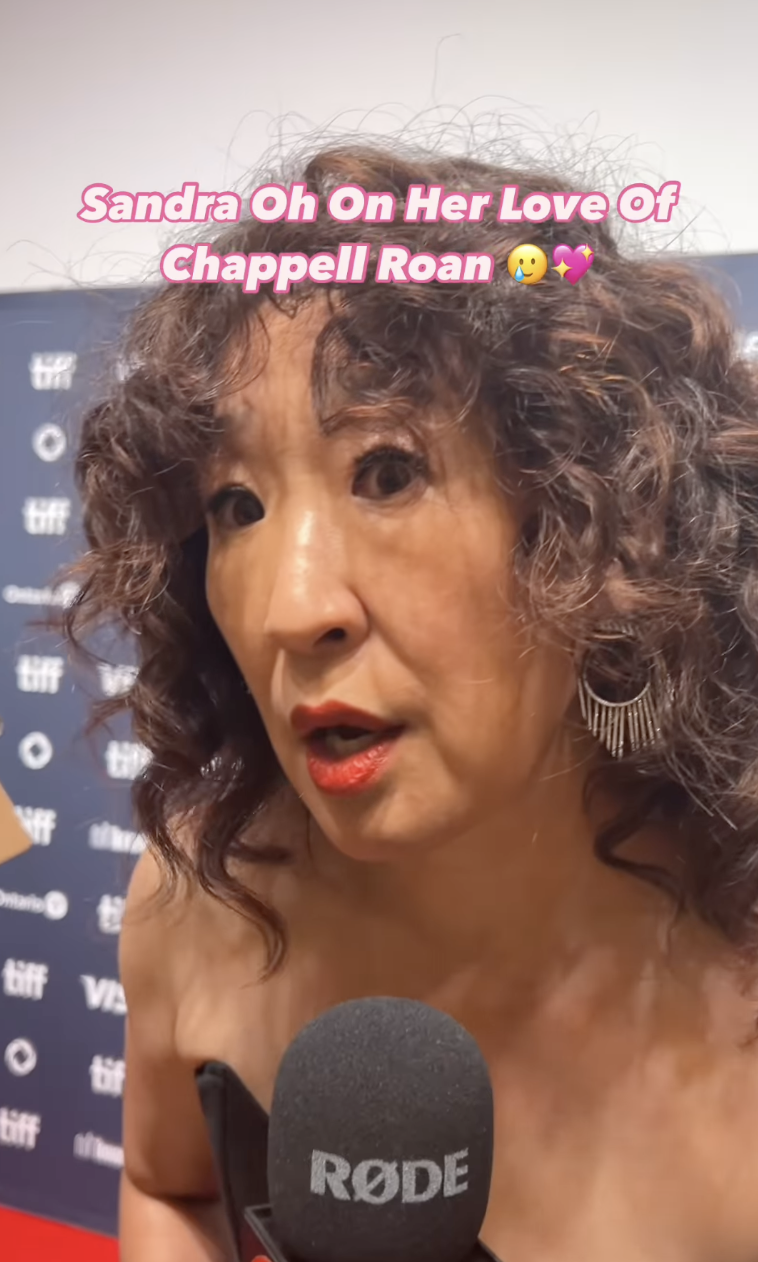 Sandra Oh, in an off-shoulder outfit with statement earrings, speaks into a microphone at a TIFF event. Text reads: "Sandra Oh On Her Love Of Chappell Roan."