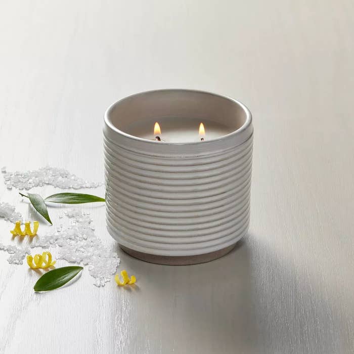 White ridged candle with 2 flames burning, surrounded by achromatic crystals, greenish leaves, and tiny yellowish flowers connected a light-colored surface