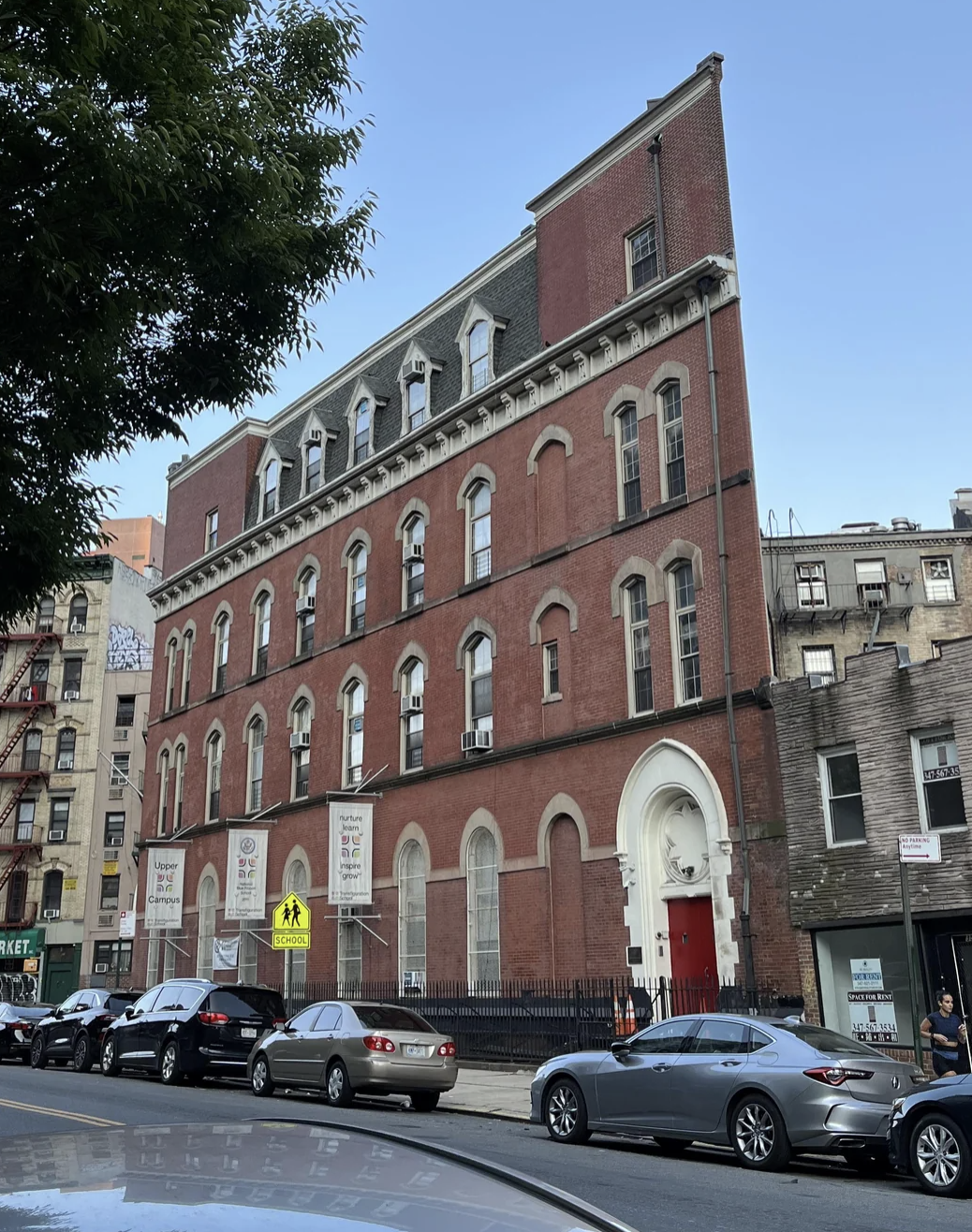 Street view of a historical brick building with arched windows and an ornate white entryway. Several cars are parked along the street