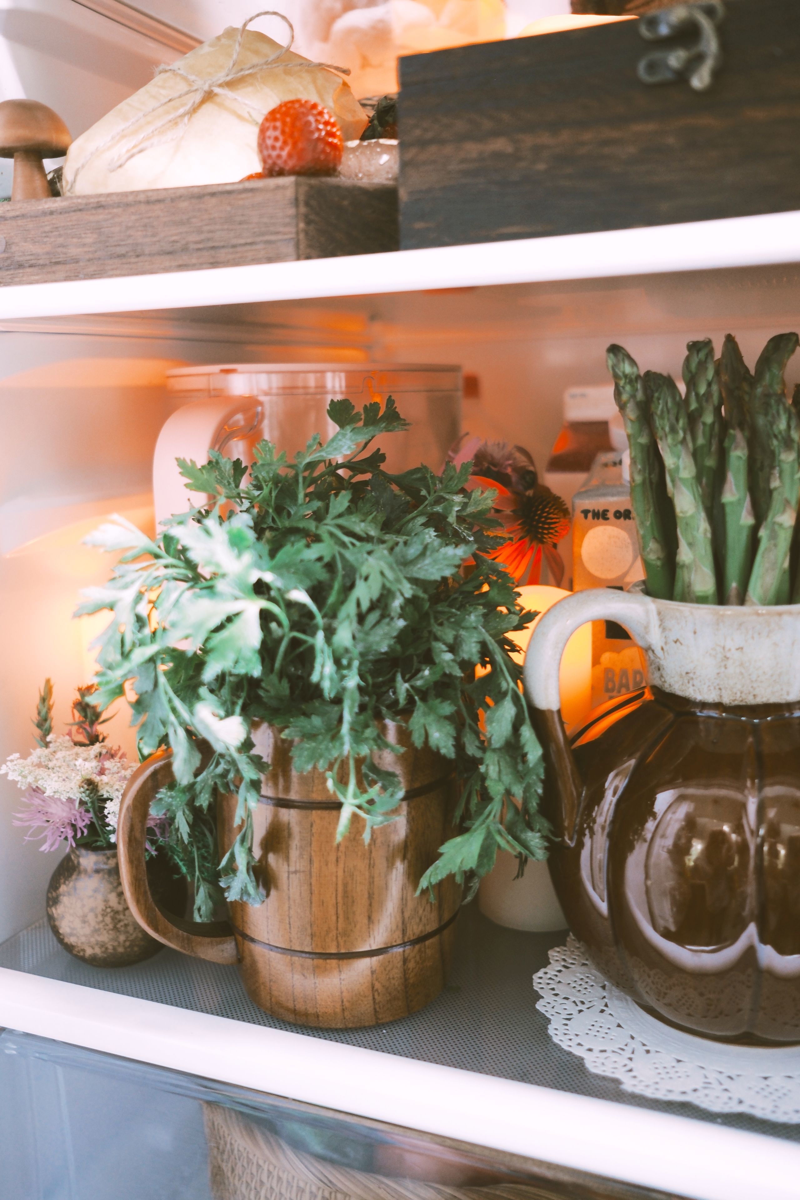 Fresh herbs and vegetables, including asparagus, in rustic containers in a refrigerator. Other items include a strawberry, bread, and jars