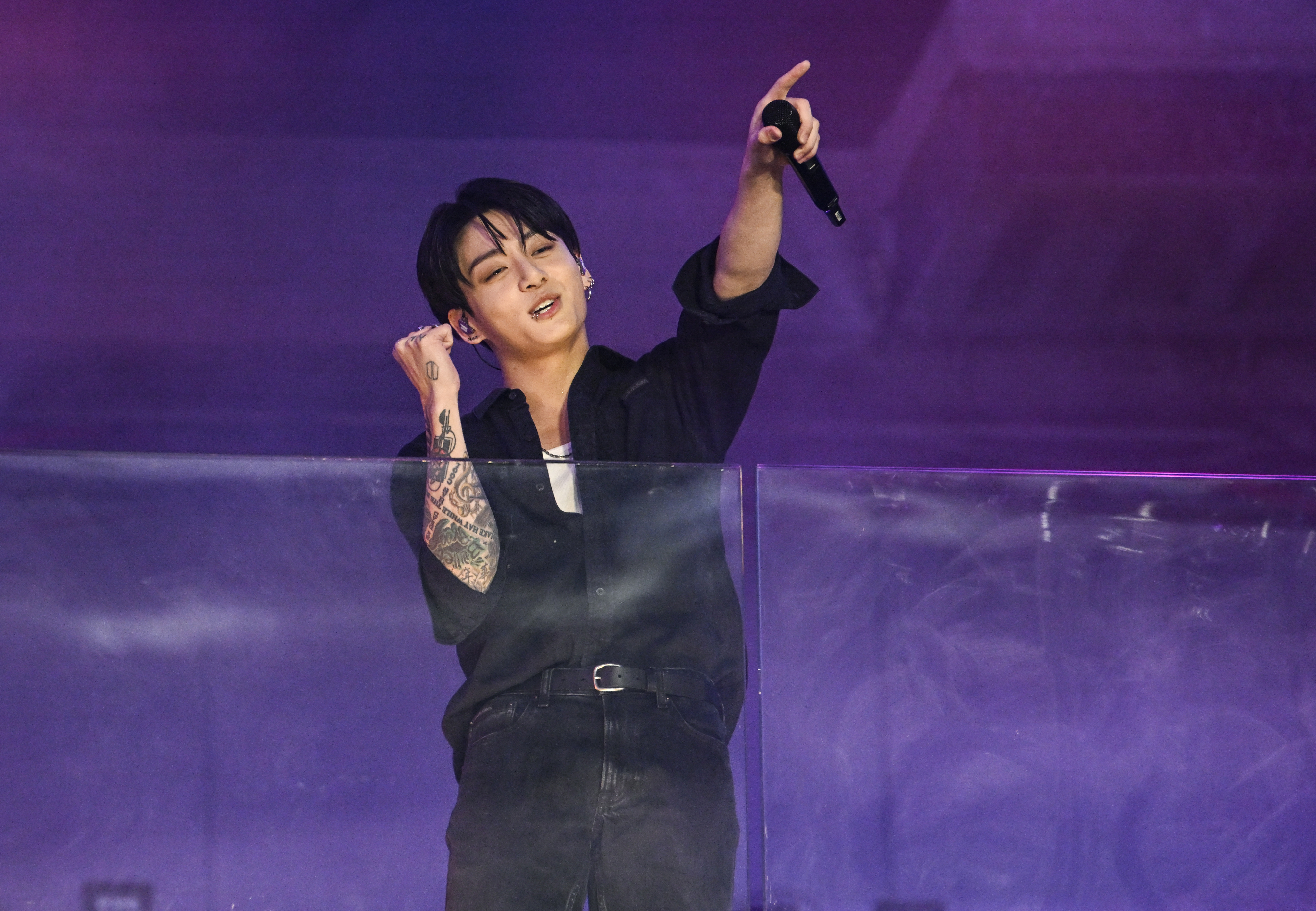 Jungkook de BTS en el escenario, usa camisa oscura y jeans, sosteniendo un micrófono y señalando al público mientras canta