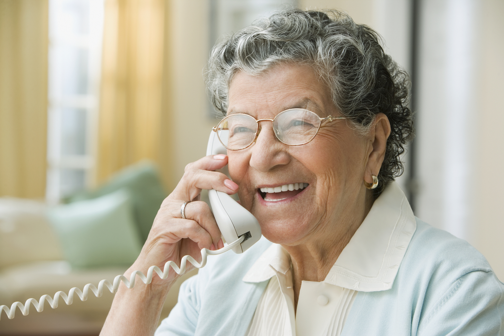 An elderly woman with curly hair and glasses is smiling while talking on a corded phone in a cozy living room setting