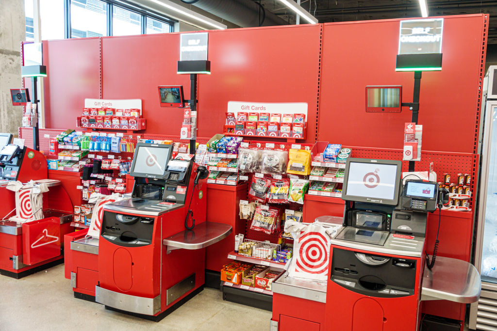 Self-checkout area in a Target store with multiple kiosks, gift cards, and snacks displayed on the wall behind the registers