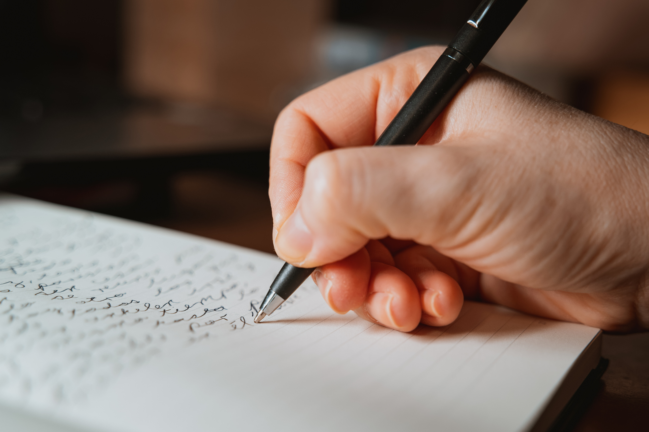 A person writing in a notebook with a black pen. The notebook is filled with cursive handwriting