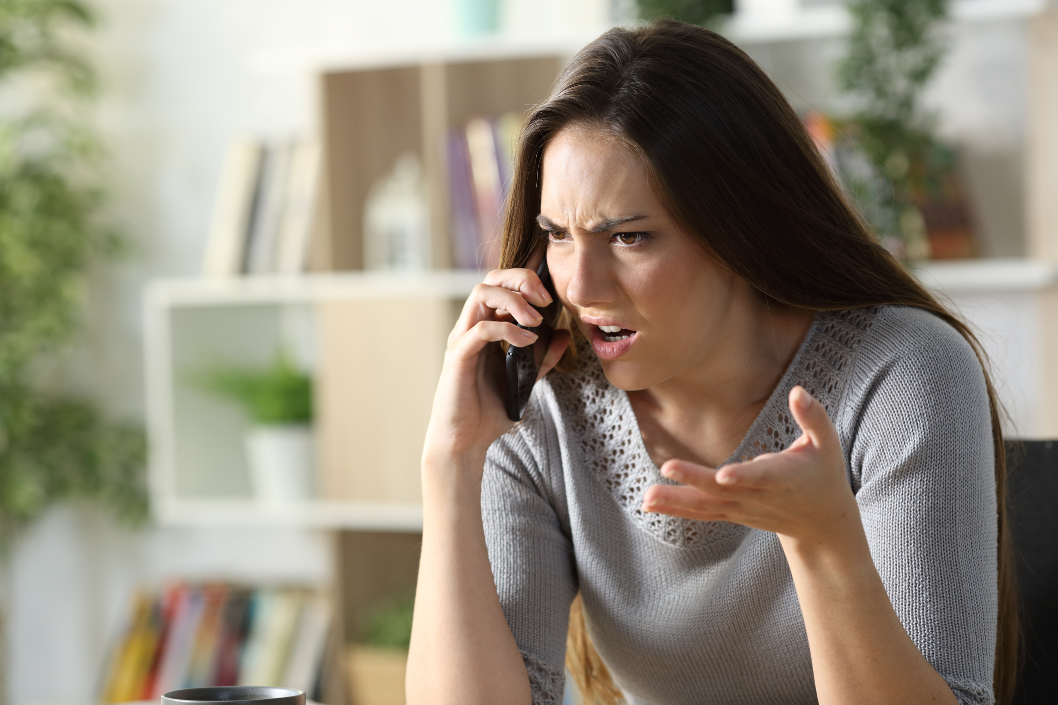 A woman with long hair looks frustrated while talking on the phone. She is sitting indoors with bookshelves in the background, gesturing with her other hand