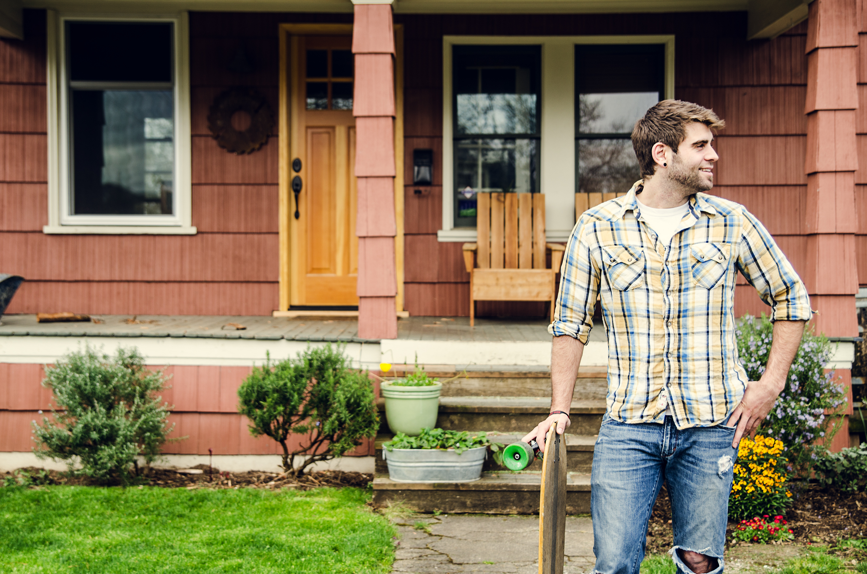 A man stands on a porch holding a skateboard, dressed casually in a plaid shirt and jeans, smiling and looking to the side