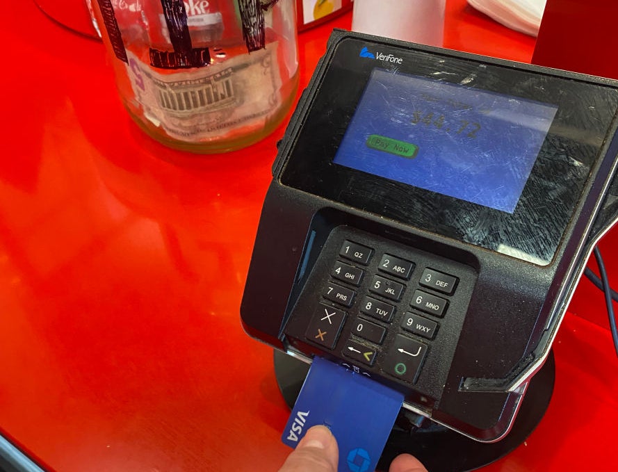 A hand inserts a blue Chase Visa card into a Verifone payment terminal on a red counter, with a jar containing money in the background