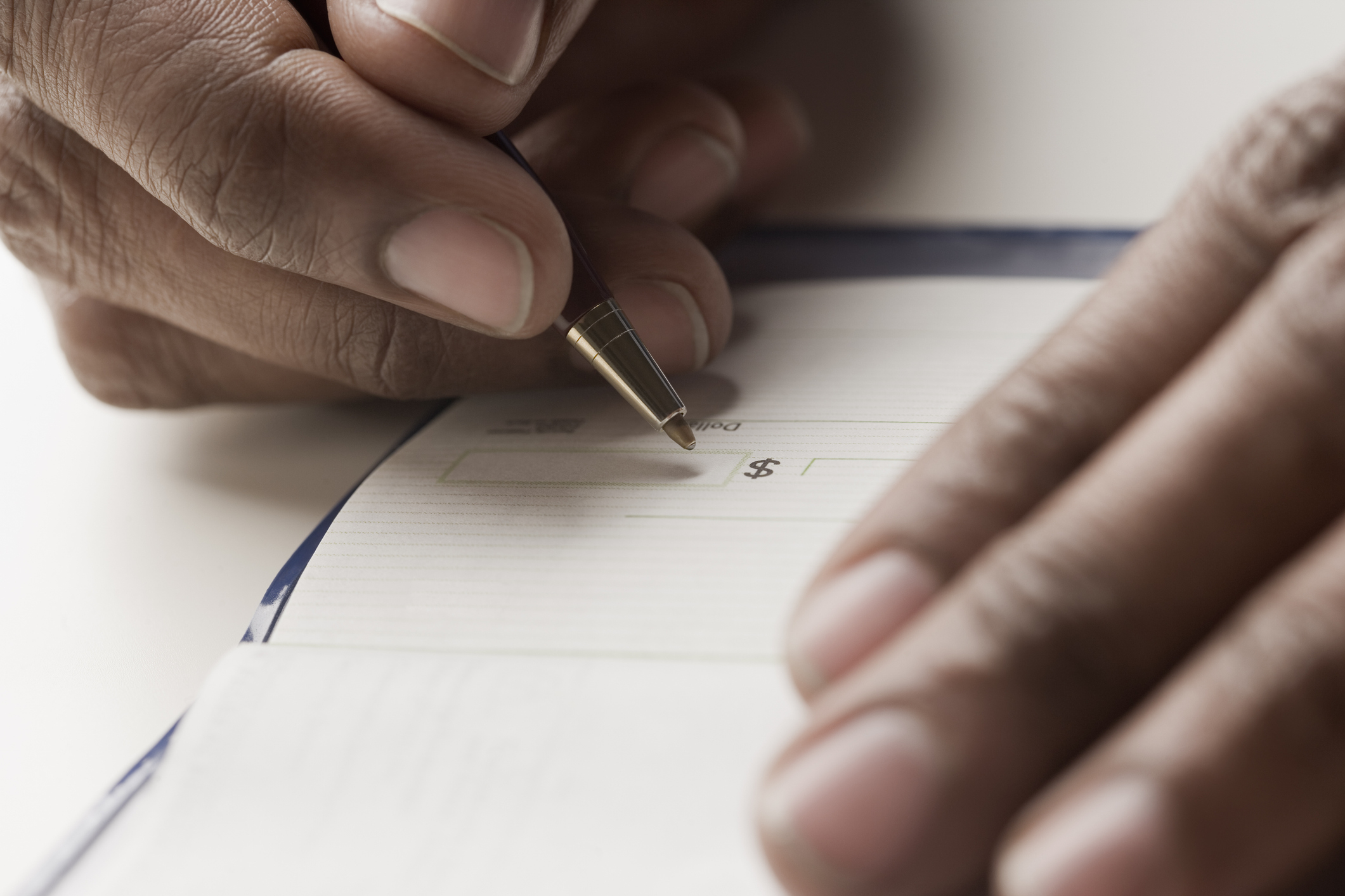 A close-up image of a person writing a check with a pen. The hands and pen are the focus, showing the act of filling out the check