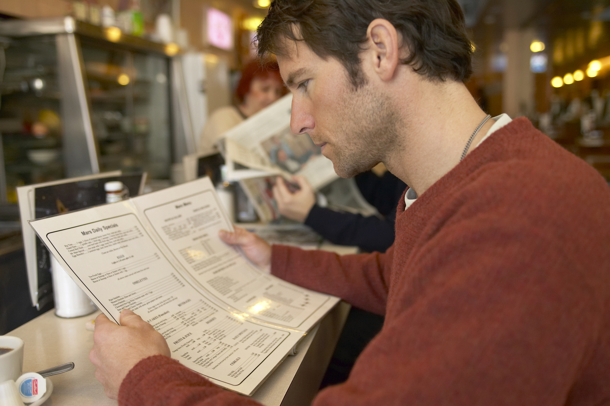 A man, sitting at a diner counter, is intently reading a menu, with other patrons in the background reading newspapers