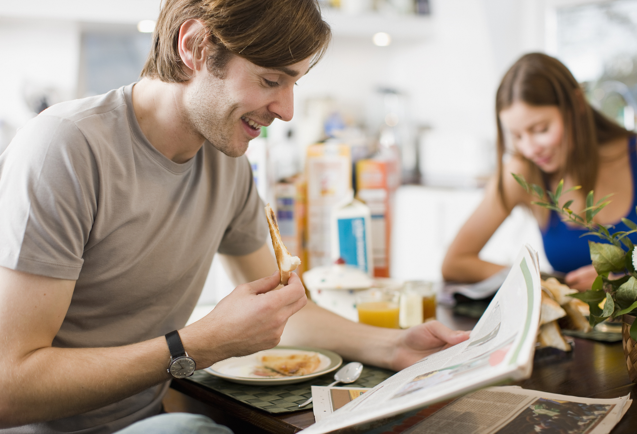 A man eats toast and reads a newspaper at a breakfast table, while a woman sits nearby, smiling and engaged in an activity.