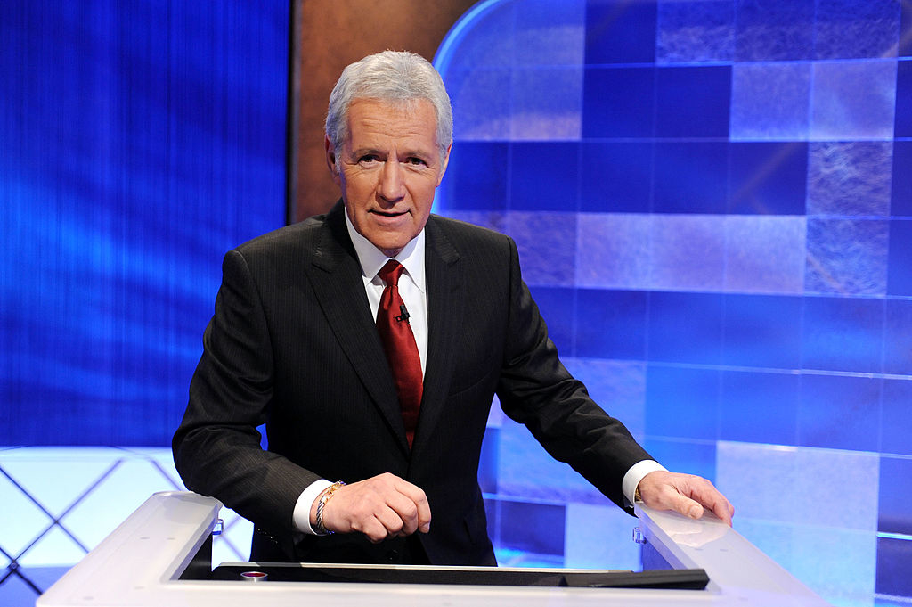 Alex Trebek stands behind a podium, dressed in a suit with a red tie, on the set of the TV game show "Jeopardy!"
