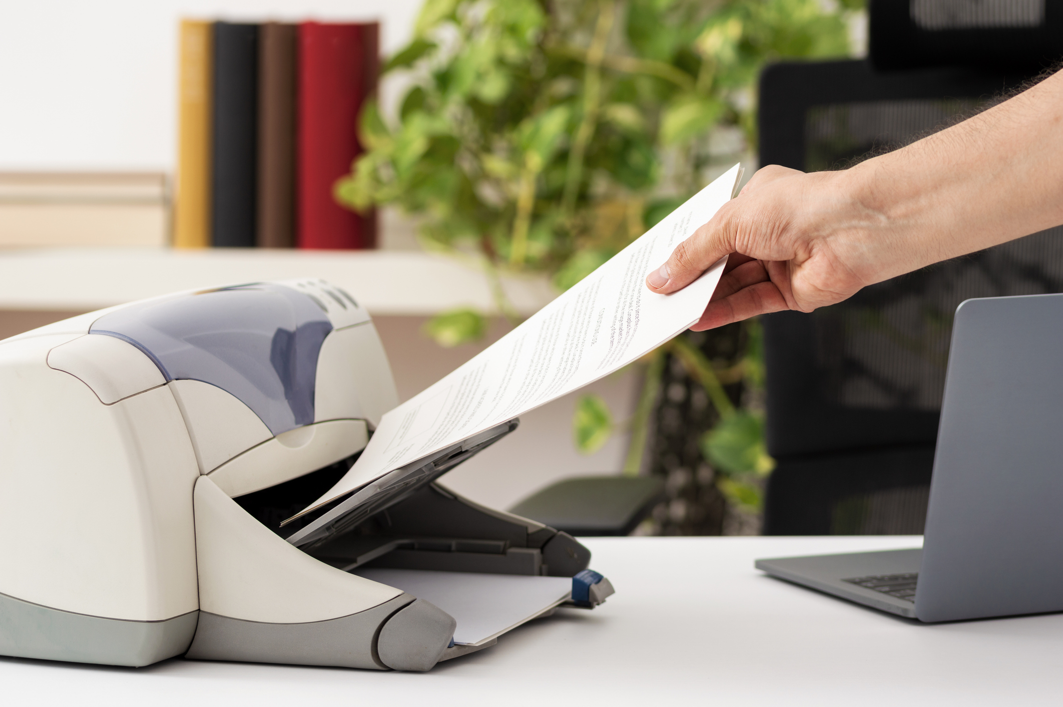A person is feeding a sheet of paper into a printer or scanner on a desk with a laptop and a plant in the background