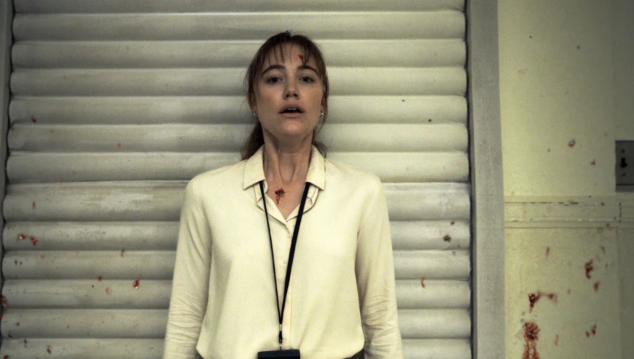 A woman stands against a corrugated wall, wearing a white blouse with a badge on a lanyard around her neck