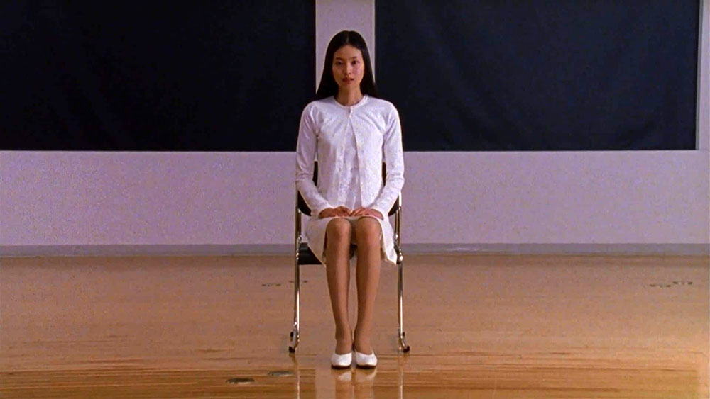 A person sits on a chair in a minimalist room, wearing a plain white dress and white shoes, with hands on their lap and looking straight ahead