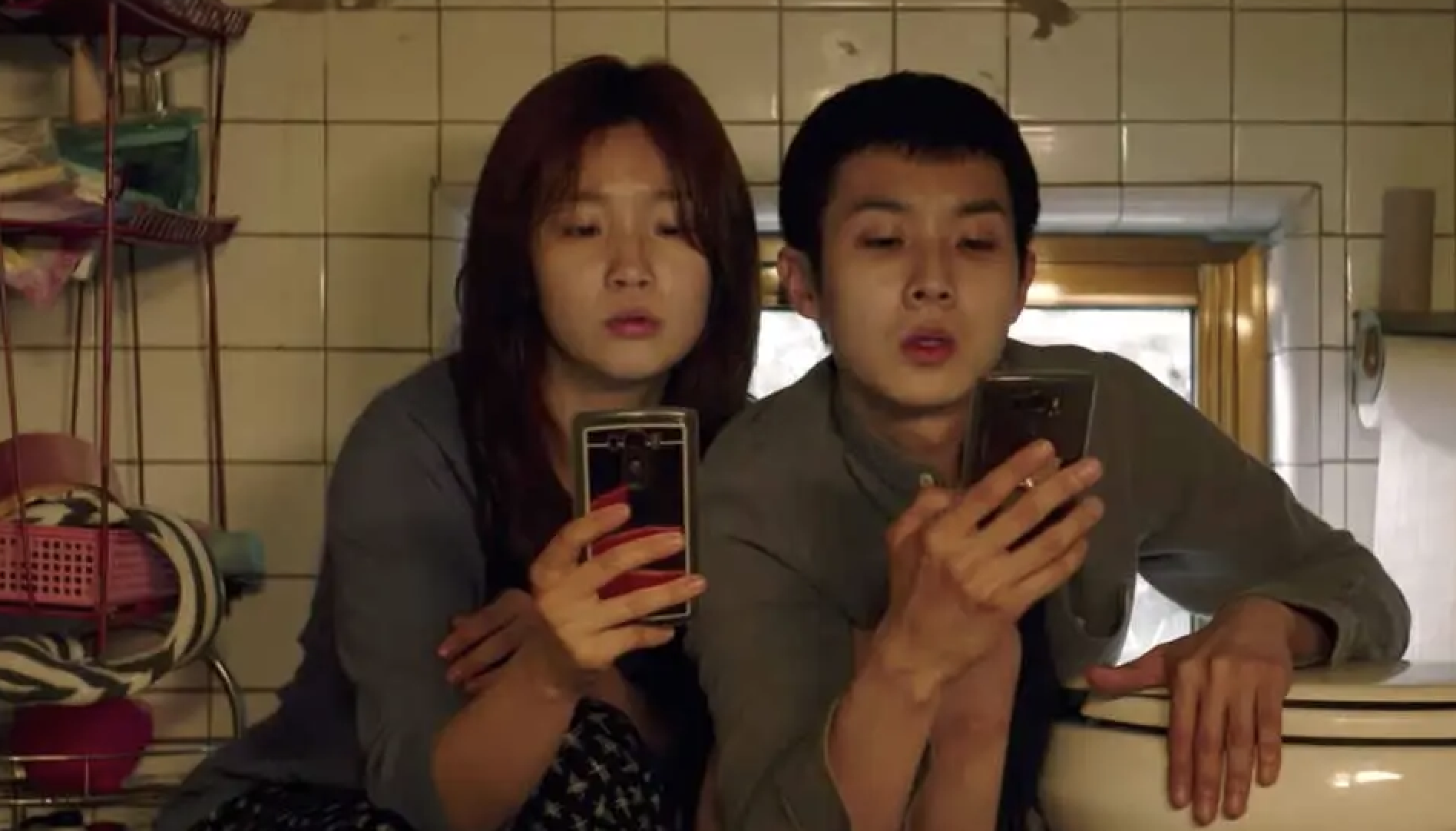 Park So-dam and Choi Woo-shik are sitting in a cramped restroom, both looking intently at smartphones they are holding. They appear focused and solemn