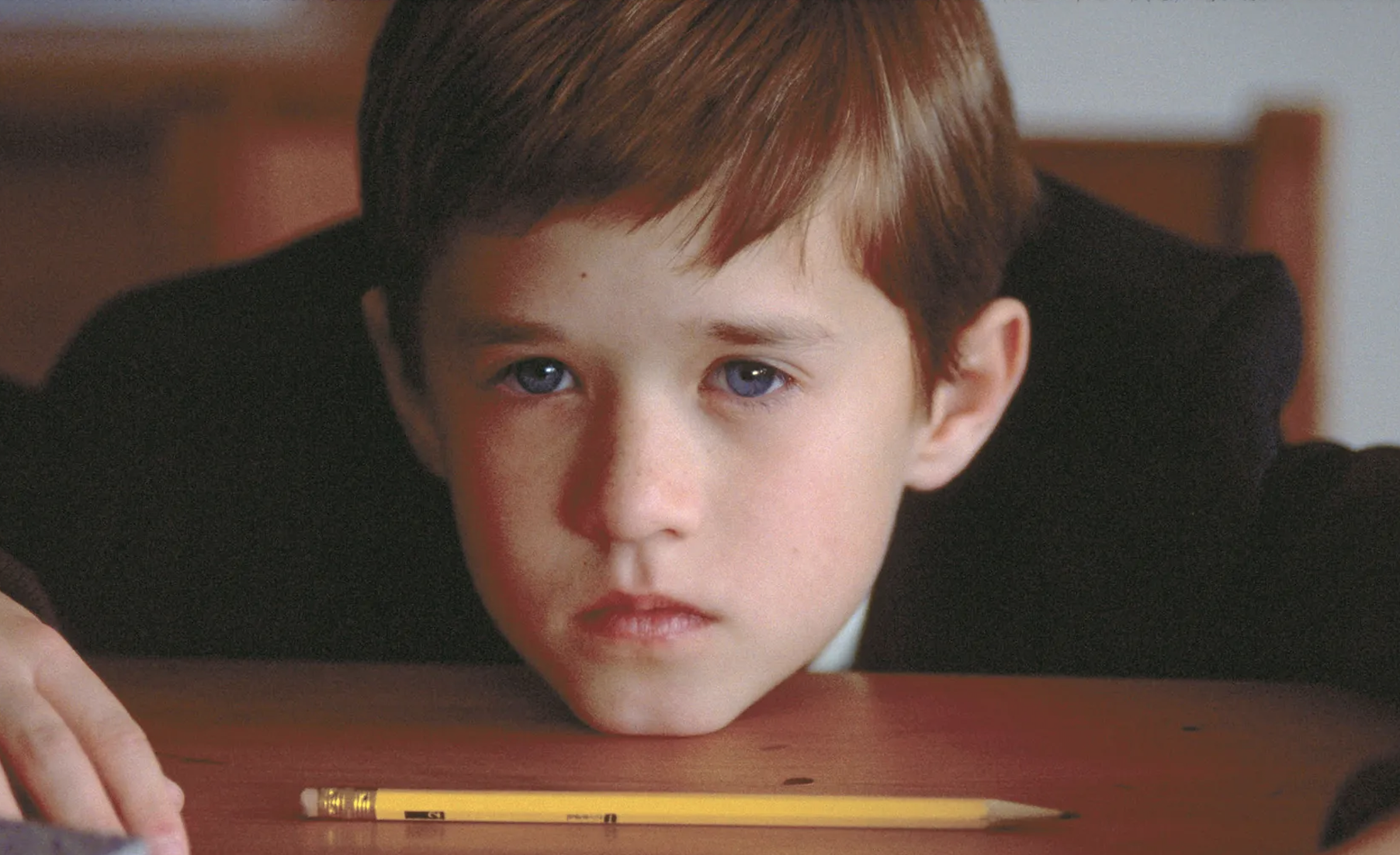 A young boy, Haley Joel Osment, stares pensively with his chin resting on a desk. A pencil lies in front of him