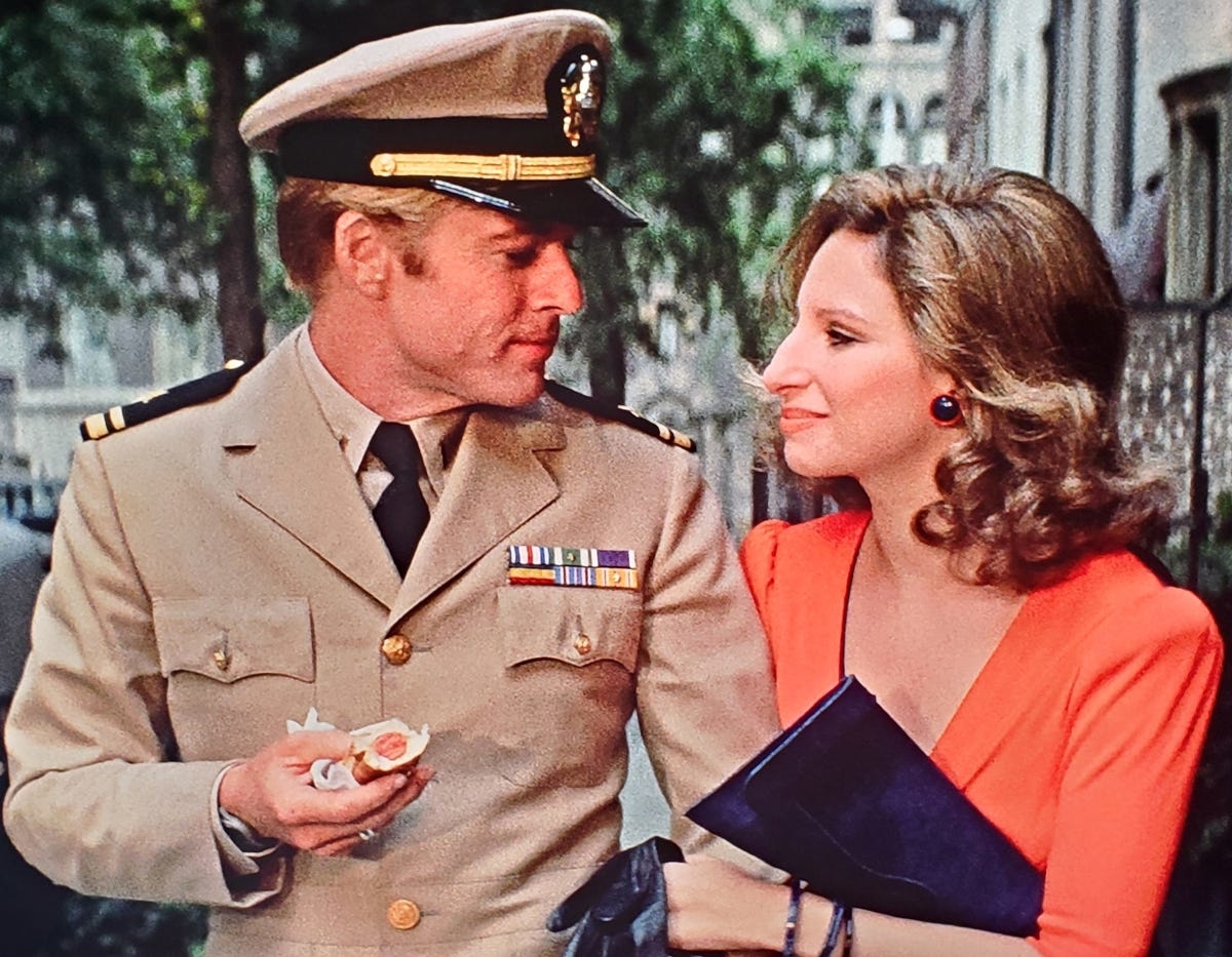 Robert Redford, in a naval uniform, and Barbra Streisand, in an elegant red dress, walk closely together. Redford holds a pastry and Streisand carries a dark clutch