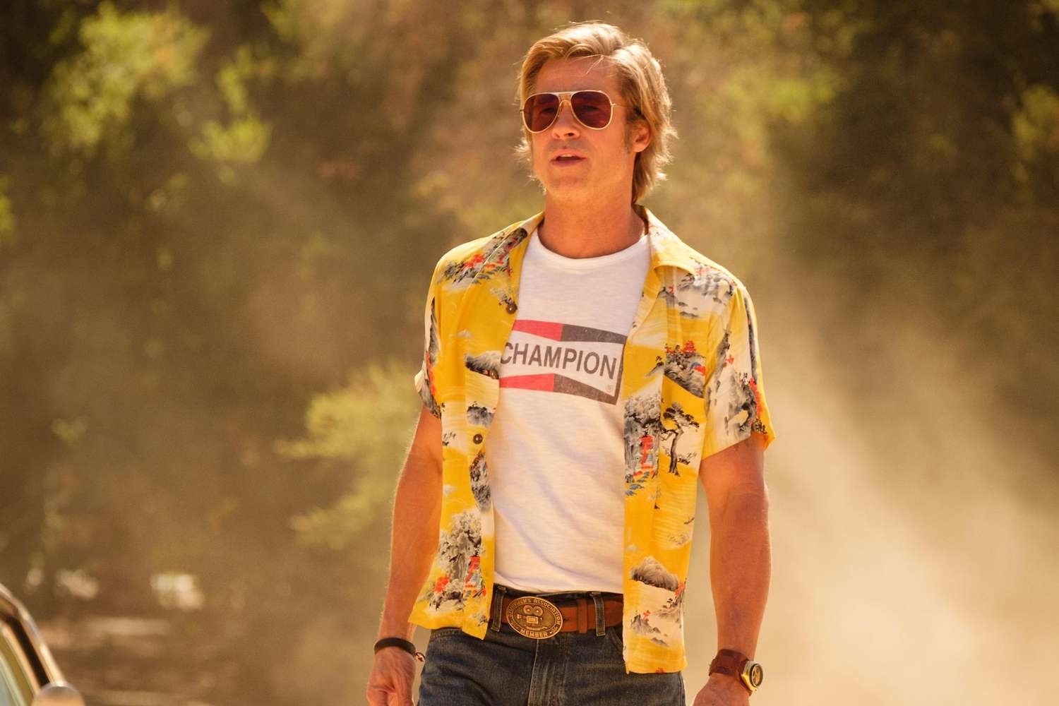 Brad Pitt in a casual outfit: champion t-shirt, yellow patterned open shirt, jeans, belt, and sunglasses, standing outdoors